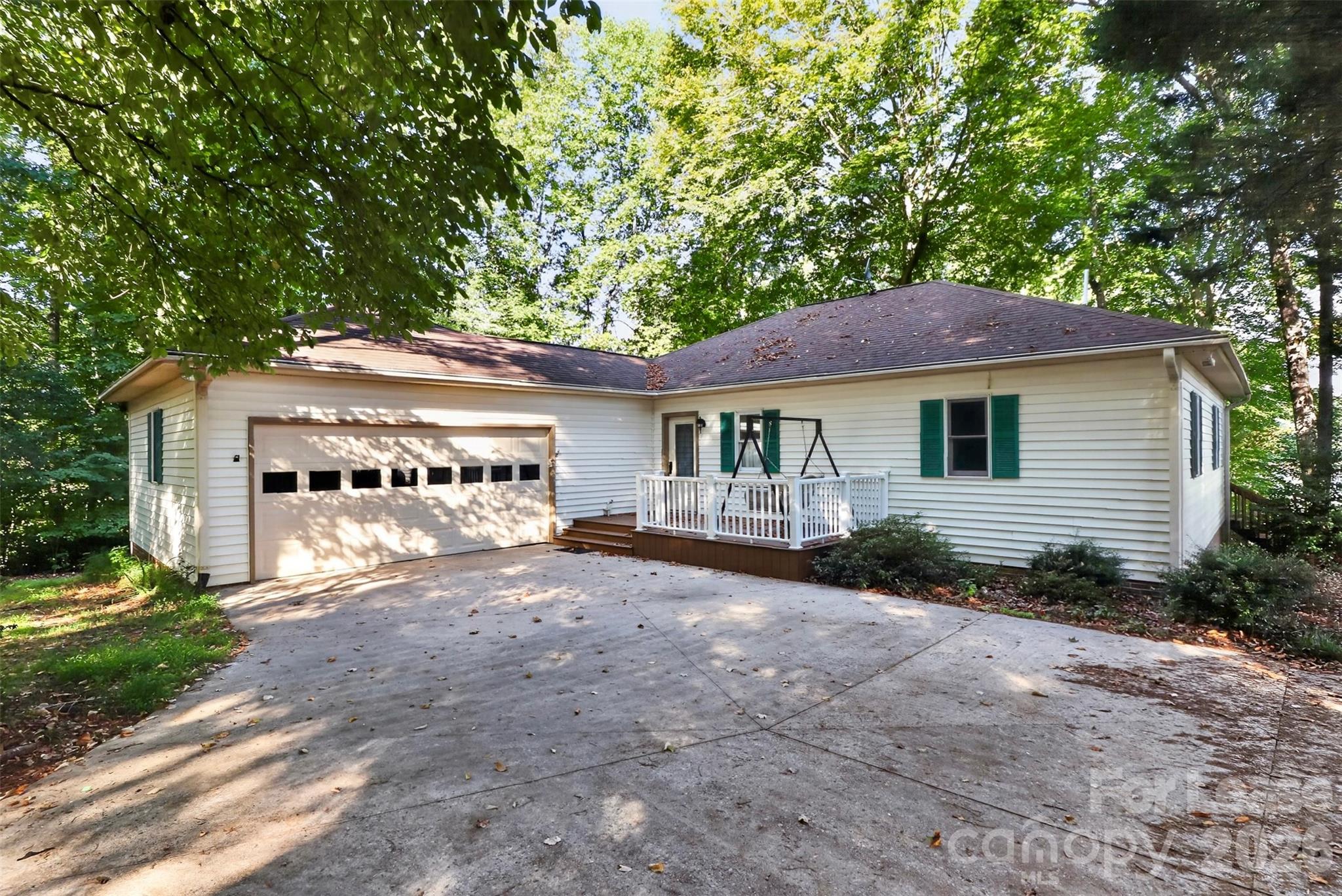 White single-story house with green shutters, a two-car garage, and a porch swing beneath mature trees.