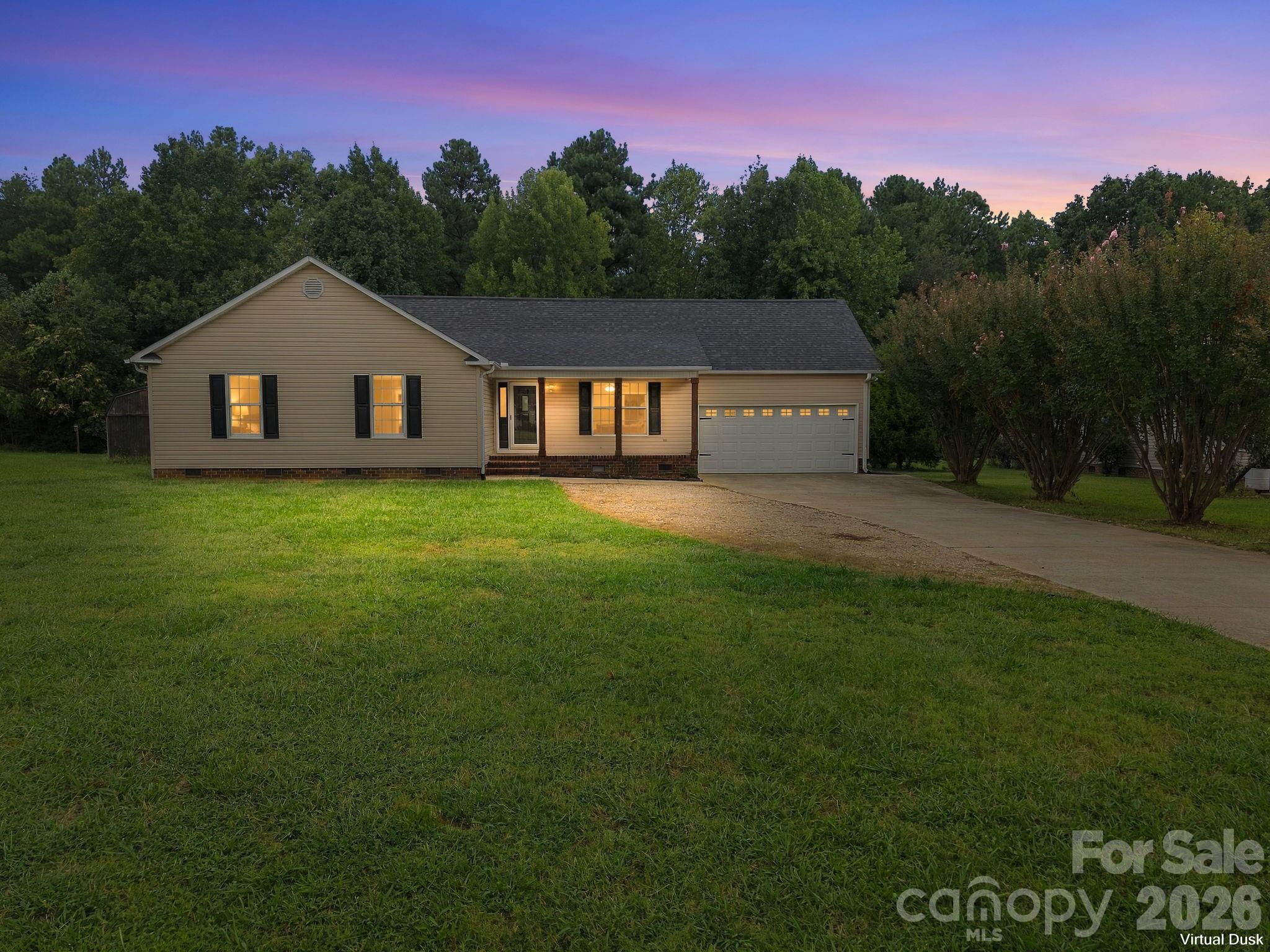 Single-story beige home with a two-car garage and glowing windows at dusk, set behind a broad lawn.