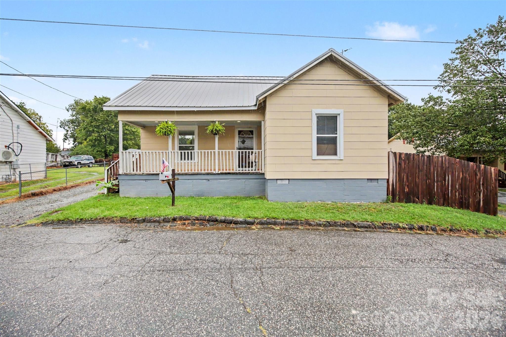 Beige house with a raised front porch, gray metal roof, and hanging plants.