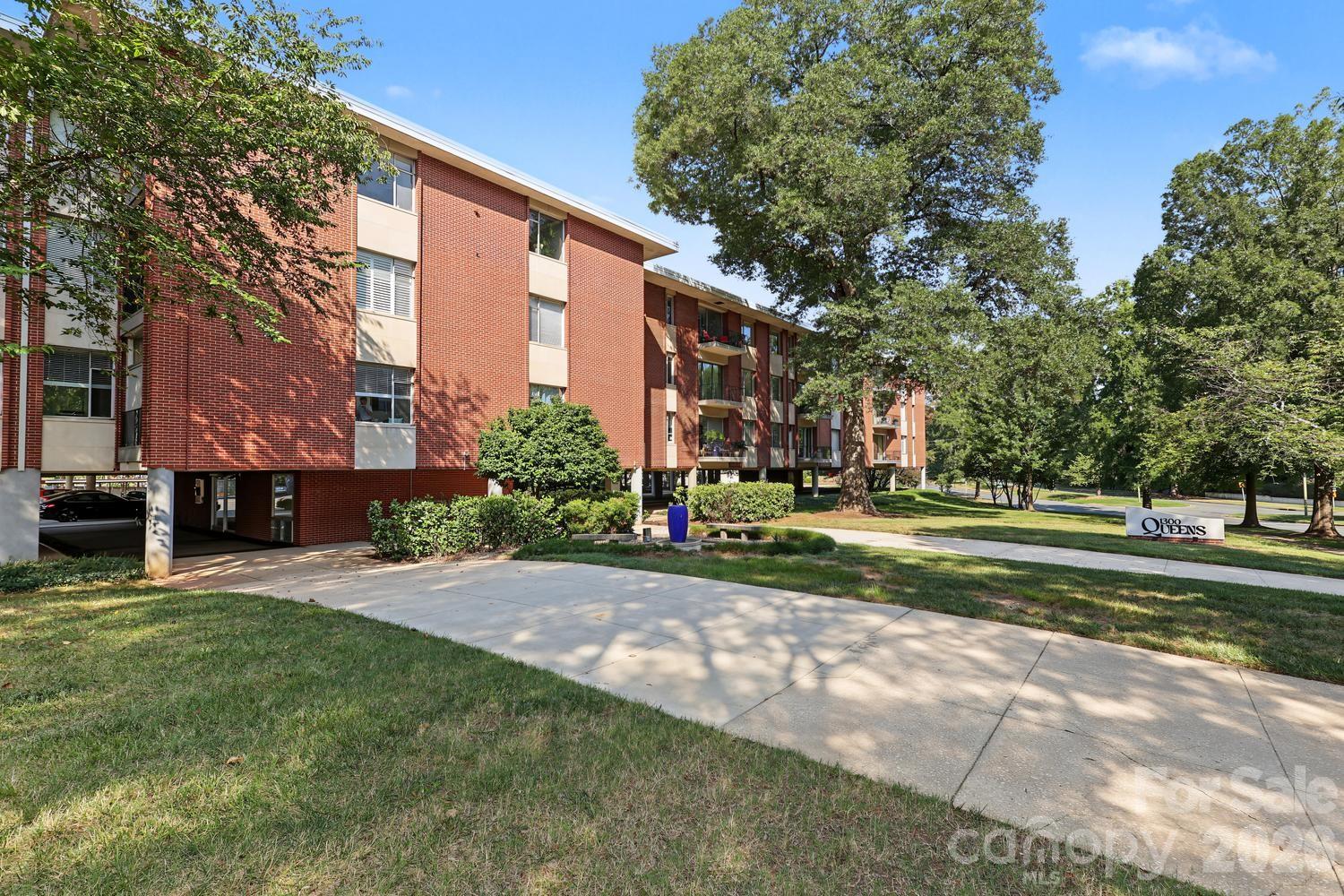 Red-brick apartment building with balconies, mature trees, and a 1300 Queens sign.