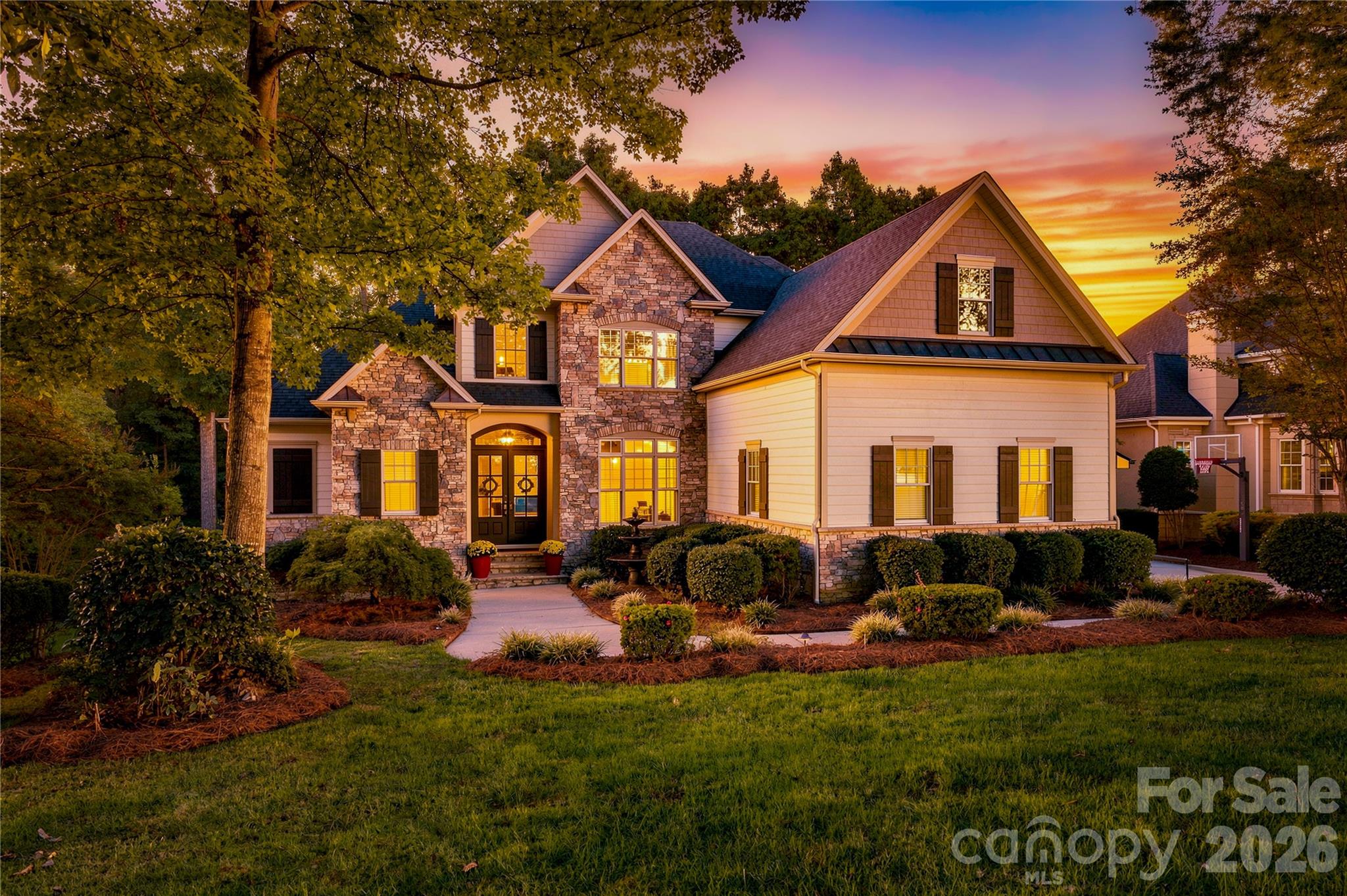 Two-story stone-and-siding house with illuminated windows and landscaped lawn at sunset.