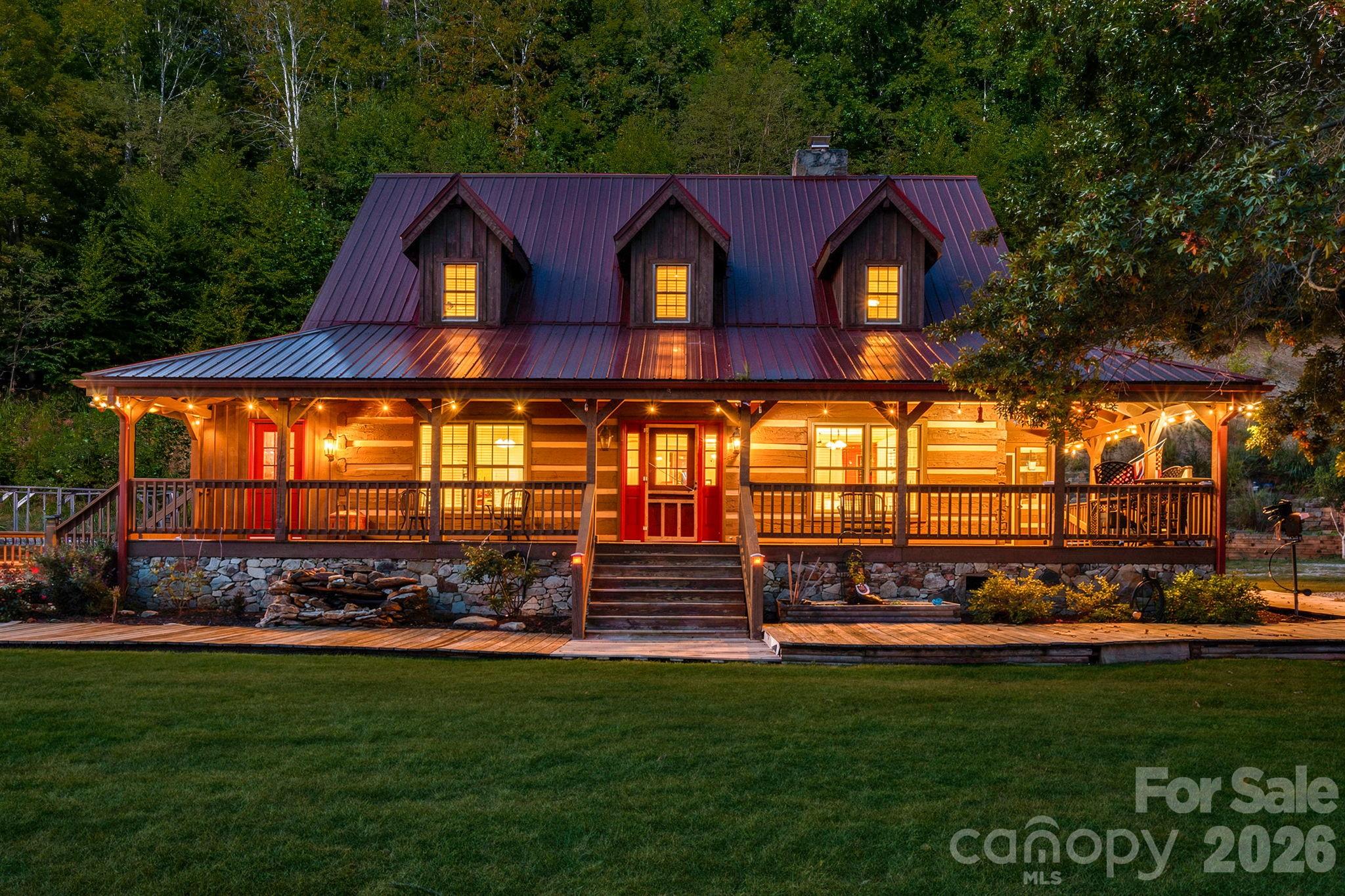 Log-style house with a burgundy metal roof and warmly lit wraparound porch at dusk.