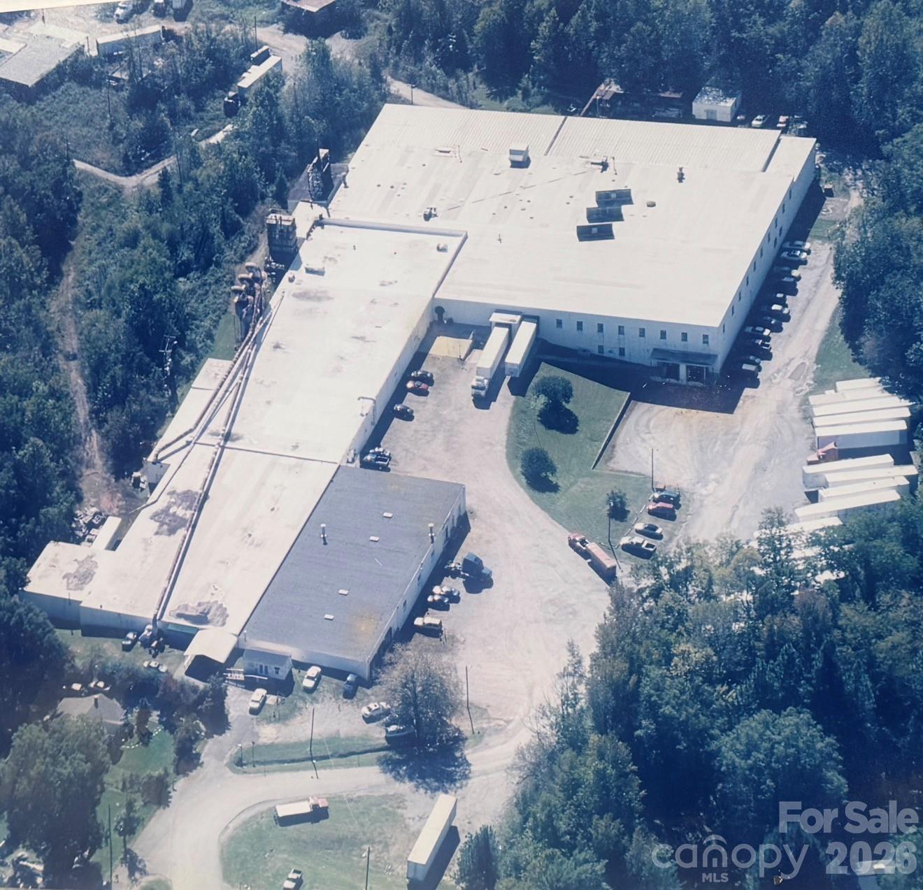 Aerial view of a large industrial building with flat roofs, parking areas, and surrounding trees.