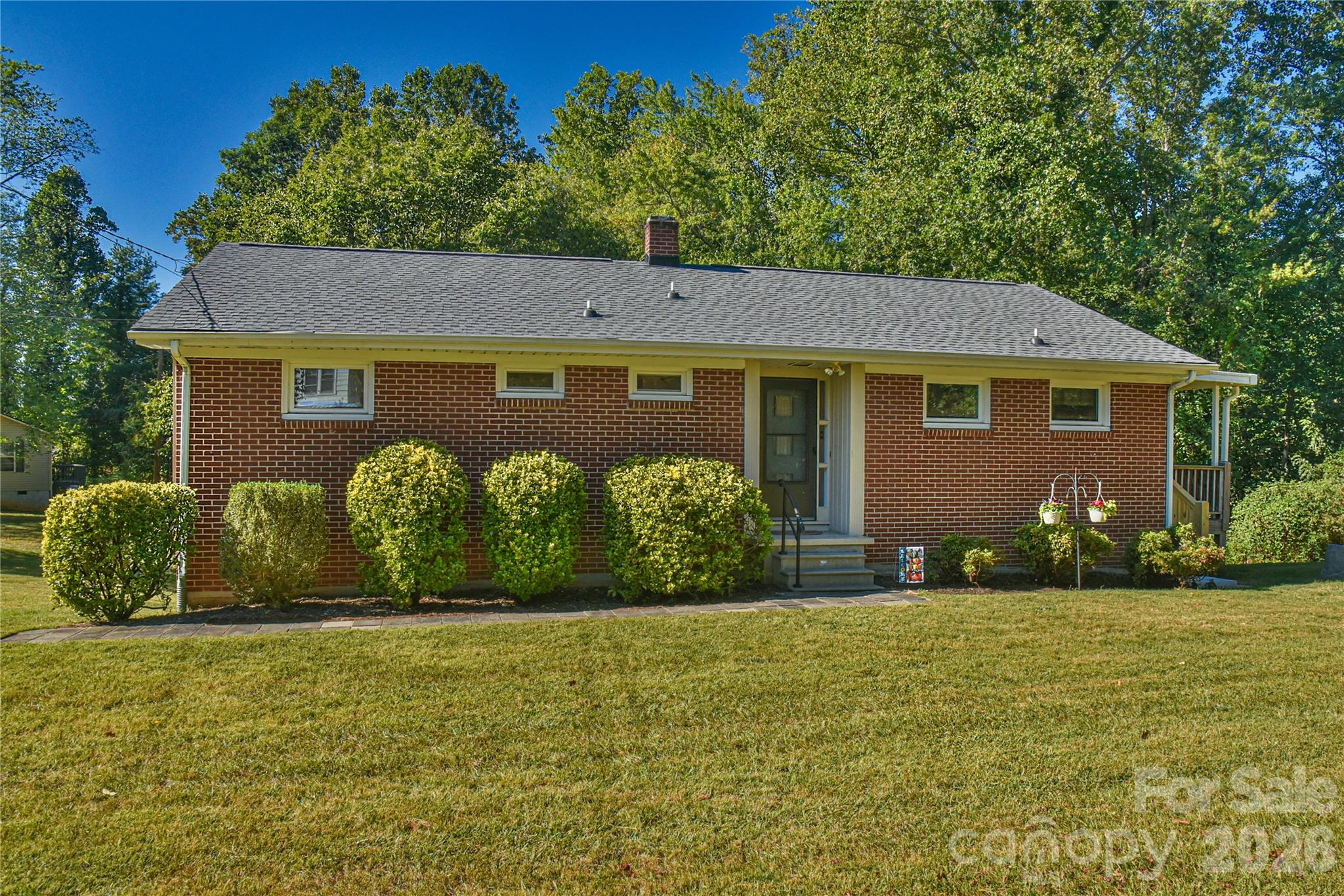 Single-story red-brick house with a gray roof, trimmed shrubs, and a broad front lawn.
