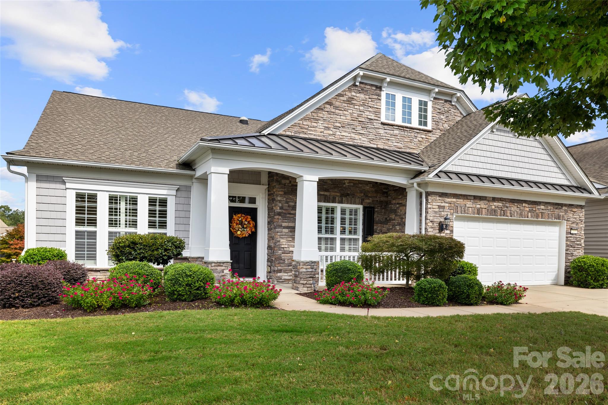 Two-story stone-and-siding house with a covered porch, white garage, and colorful landscaped flower beds.