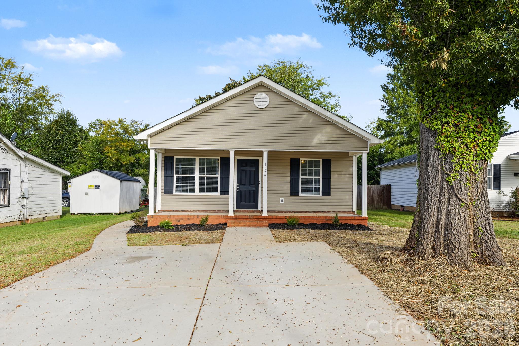 Beige house with a covered porch and wide driveway, framed by a large ivy-covered tree.