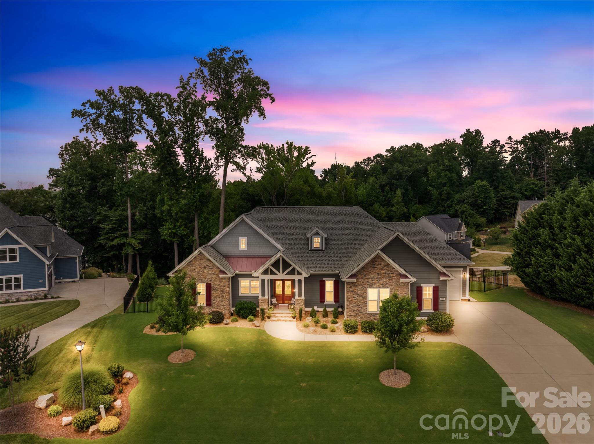 Aerial view of a stone-and-gray house with red shutters, landscaped lawn, and mature trees at dusk.