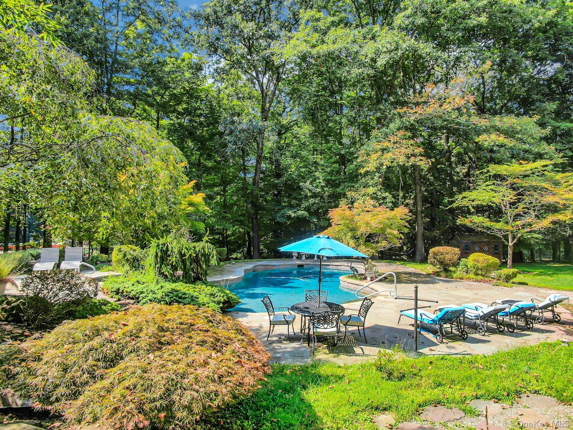 Backyard swimming pool with a turquoise umbrella, stone patio, and lounge chairs beneath mature trees.