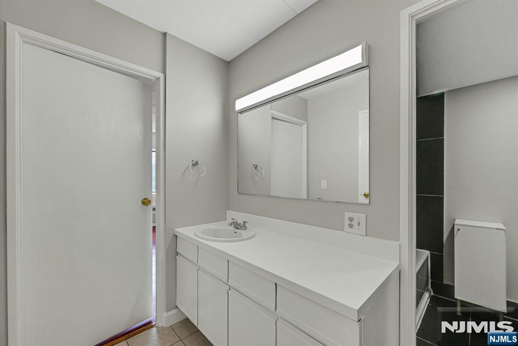 Bathroom with a long white vanity, single sink, and illuminated mirror beside a dark-tiled shower.