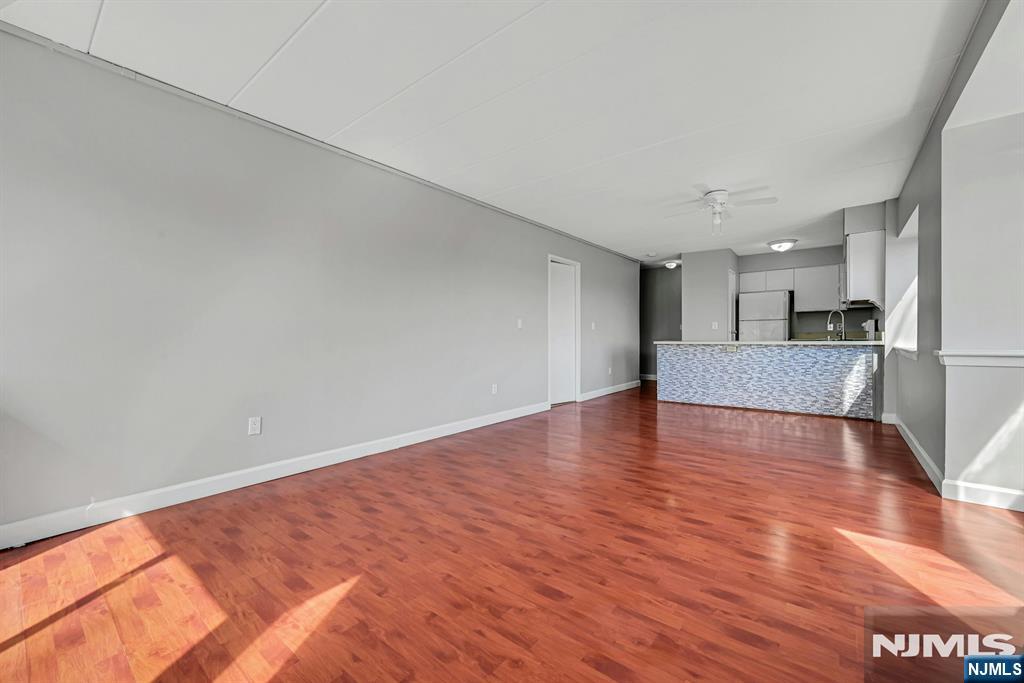 Empty living area with reddish hardwood floors opening to a kitchen with a tiled peninsula counter.