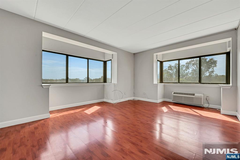 Empty room with reddish hardwood floors, two wide windows, and a wall-mounted air conditioner.