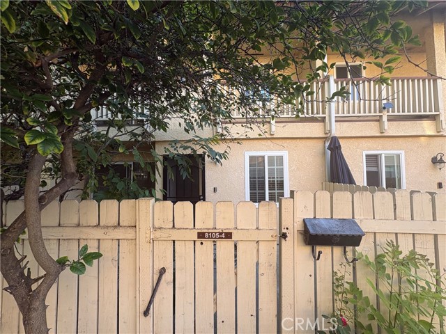 Townhouse exterior behind a wooden fence, with a second-floor balcony and leafy tree.