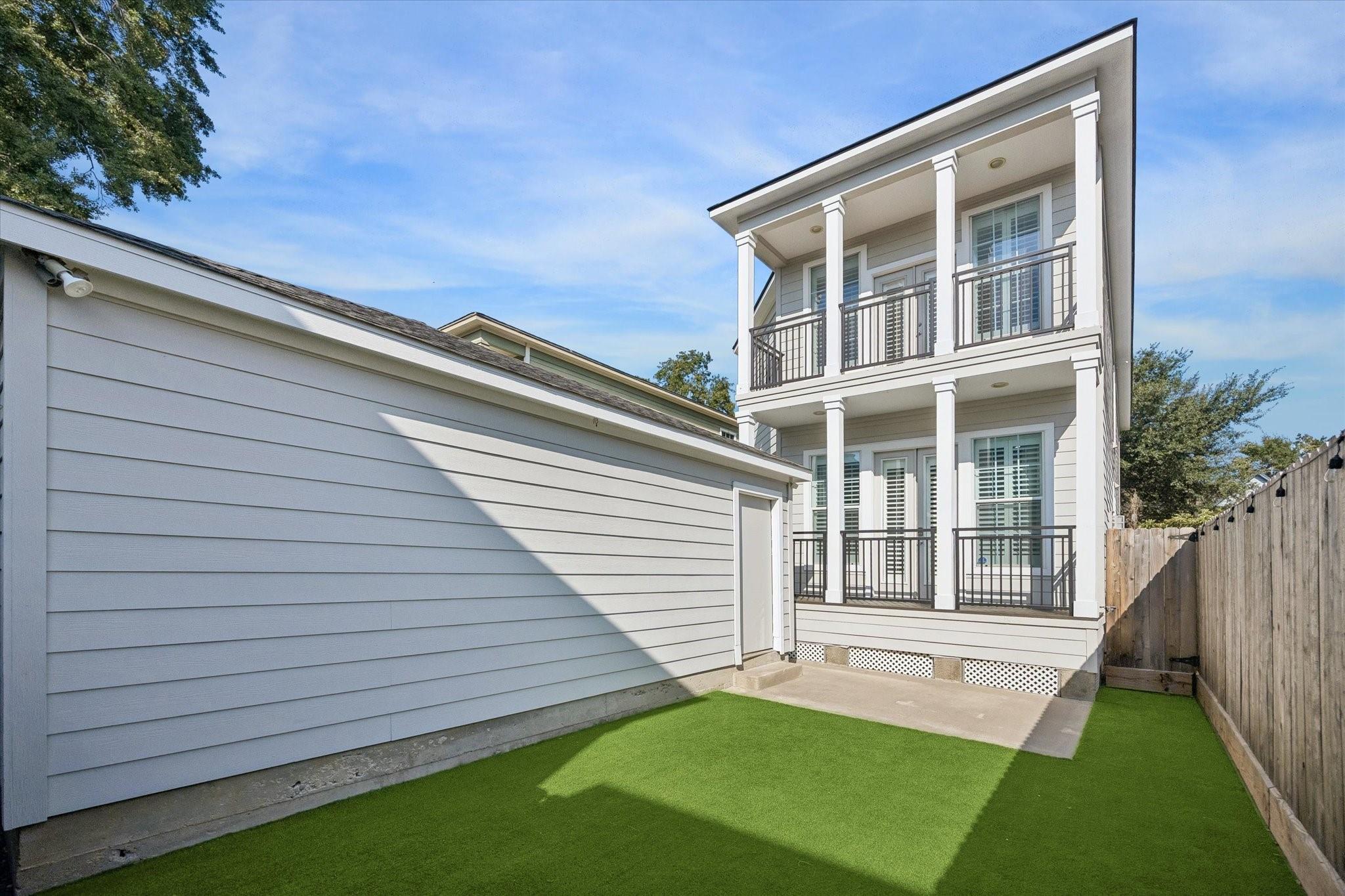 Rear of a two-story house with stacked balconies, white columns, and a fenced artificial-turf yard.