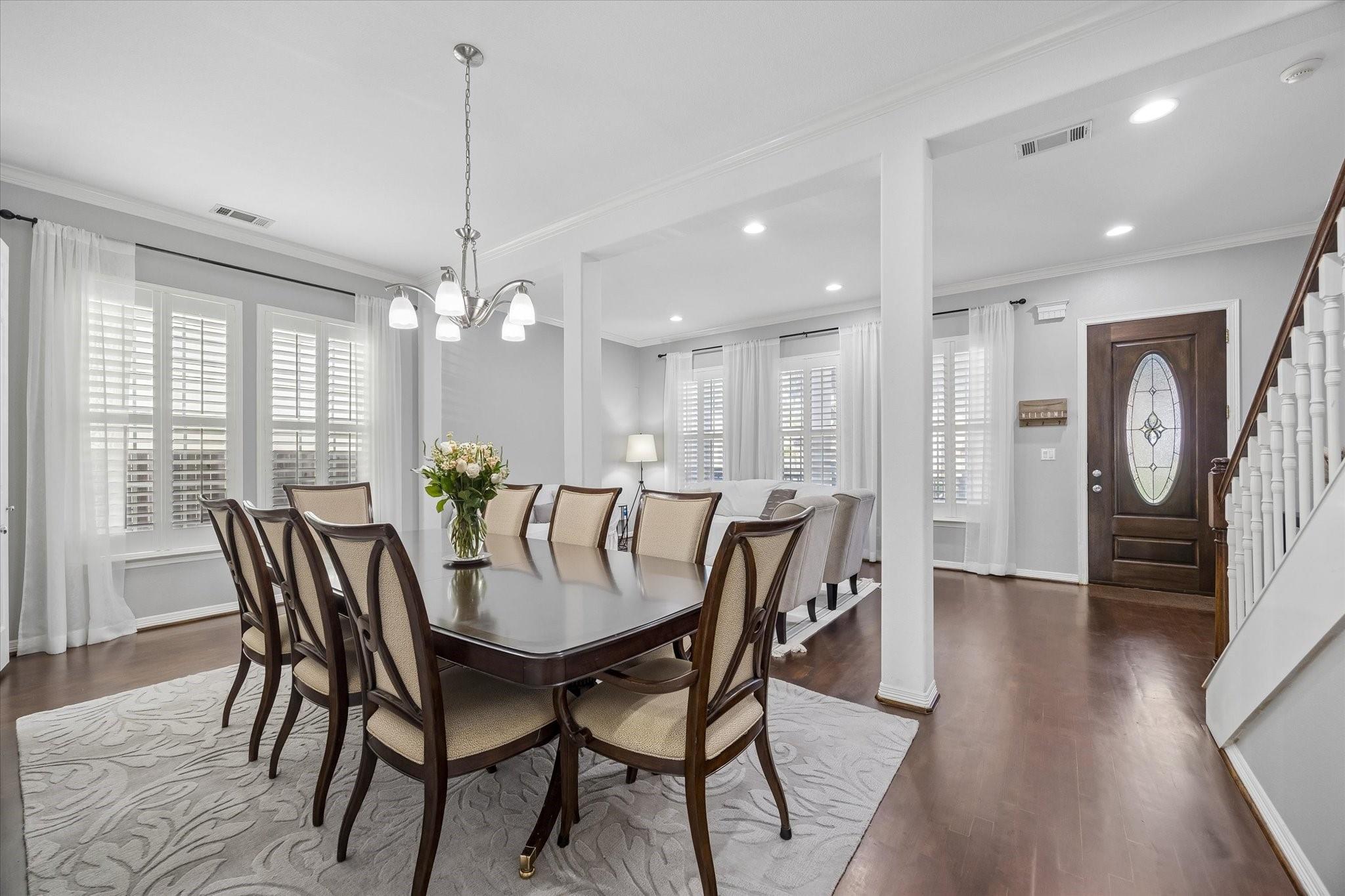 Dining room with a long table, upholstered chairs, shuttered windows, and an open view toward the living area.