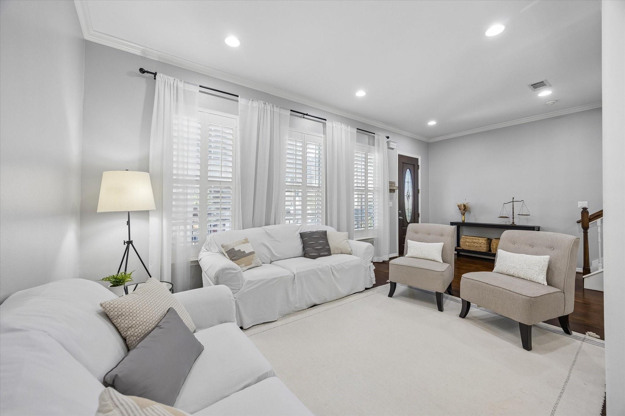 Living room with white sofas, two upholstered chairs, plantation shutters, and a bright neutral palette.