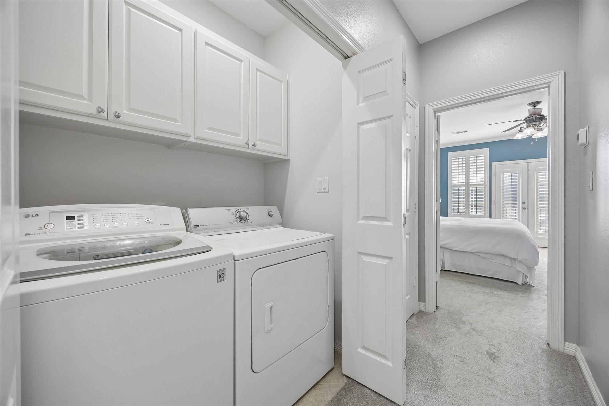 Laundry room with washer and dryer beneath white cabinets, opening to a bedroom.