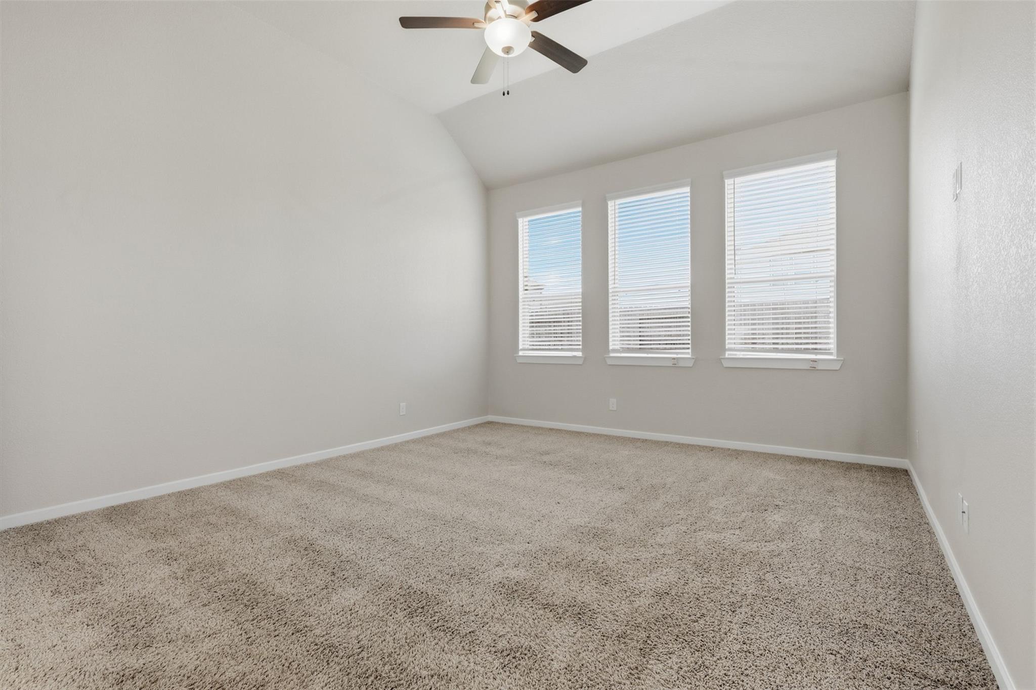 Empty bedroom with beige carpet, vaulted ceiling, ceiling fan, and three windows with blinds.
