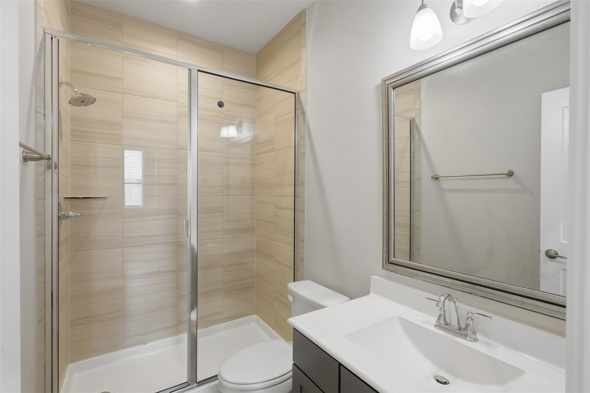 Bathroom with a beige-tiled glass shower, white toilet, and dark vanity with a large mirror.