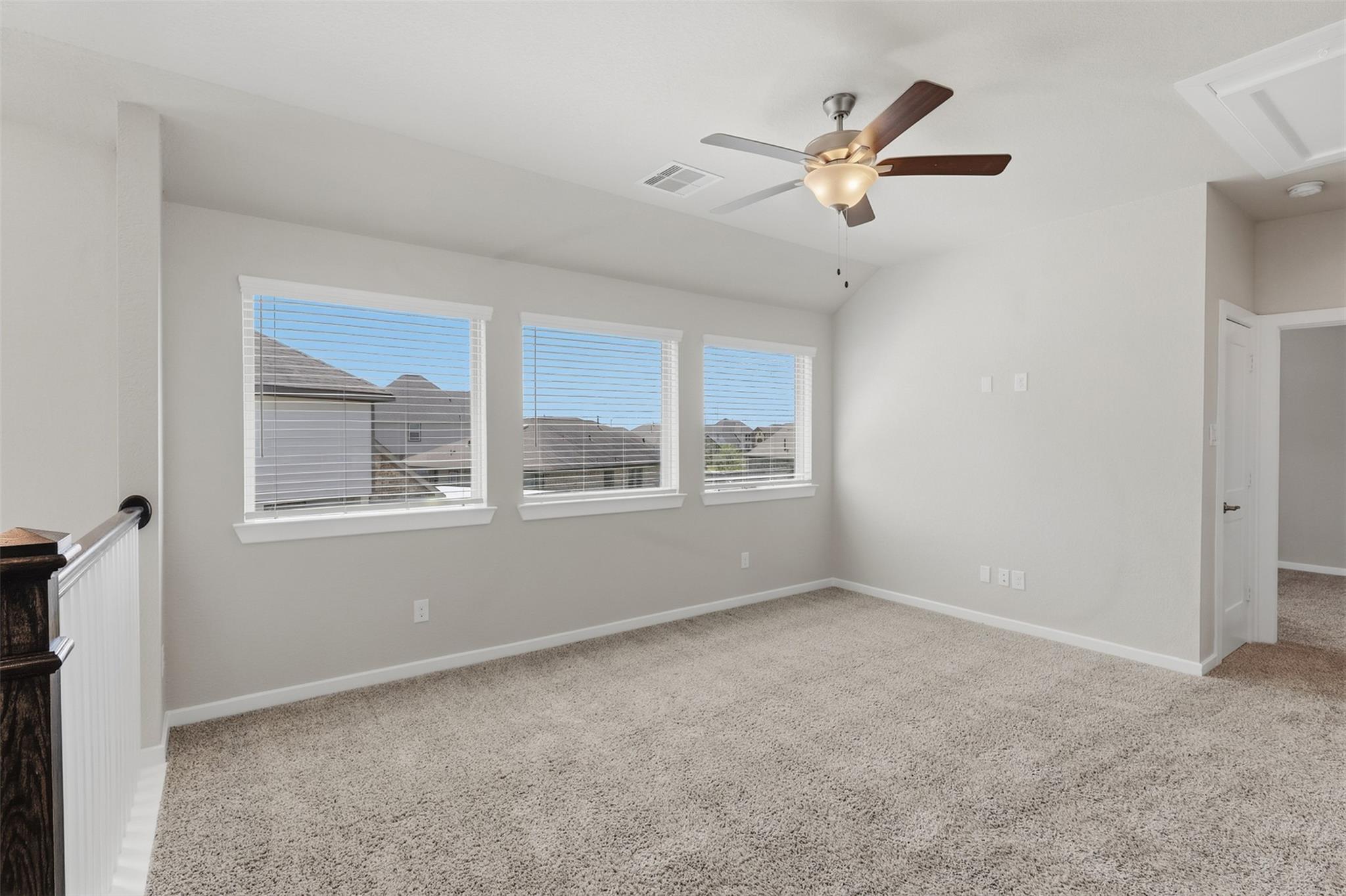 Empty carpeted upstairs room with three windows, a ceiling fan, and a stair railing.