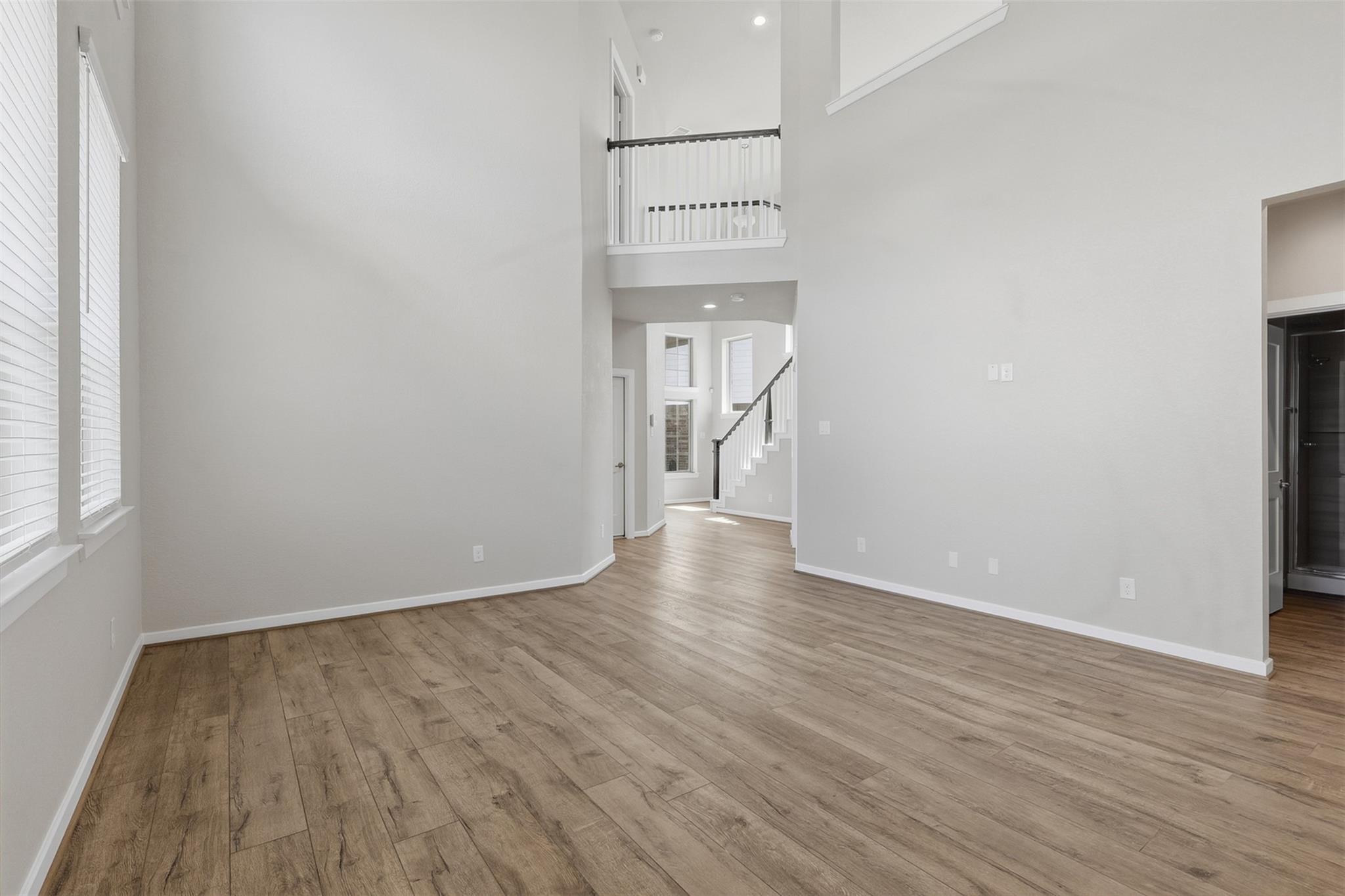 Empty living area with wood-look floors, tall windows, and a staircase leading to an upstairs landing.