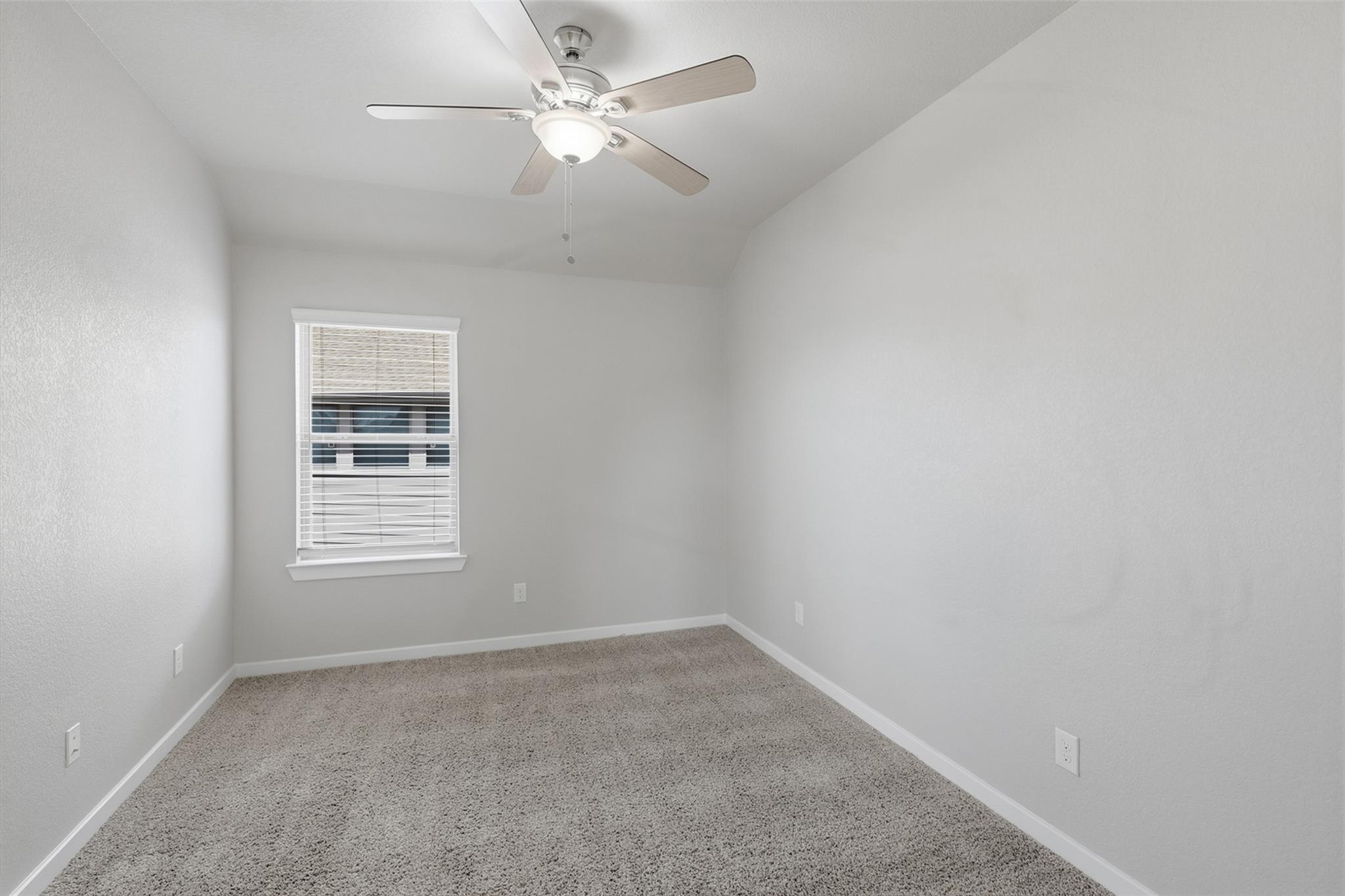 Empty bedroom with beige carpet, vaulted ceiling, ceiling fan, and window with blinds.