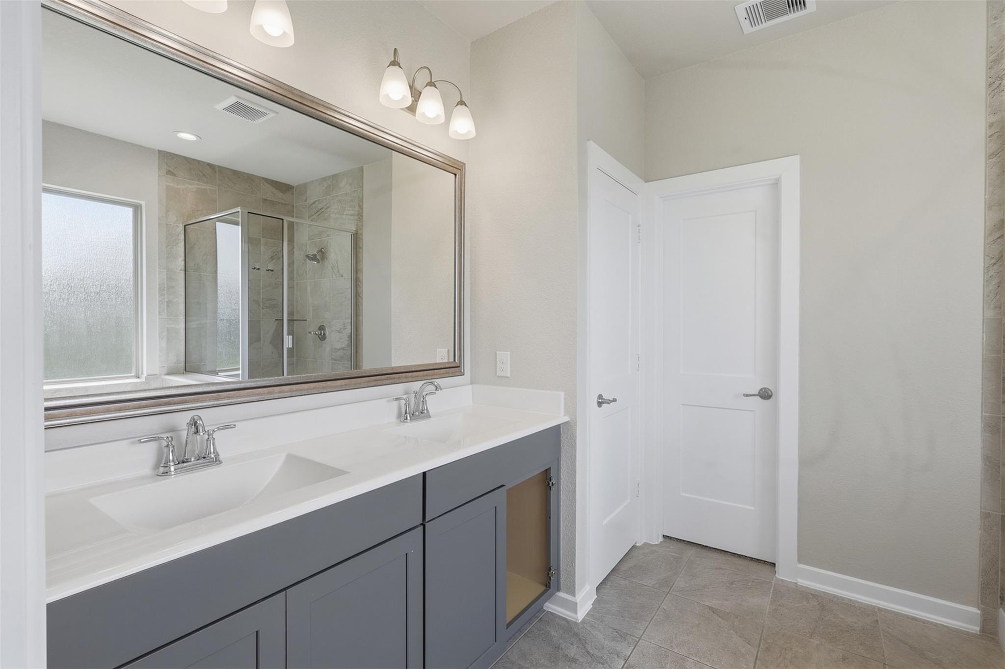 Bathroom with a gray double-sink vanity and a large mirror reflecting a tiled glass shower.