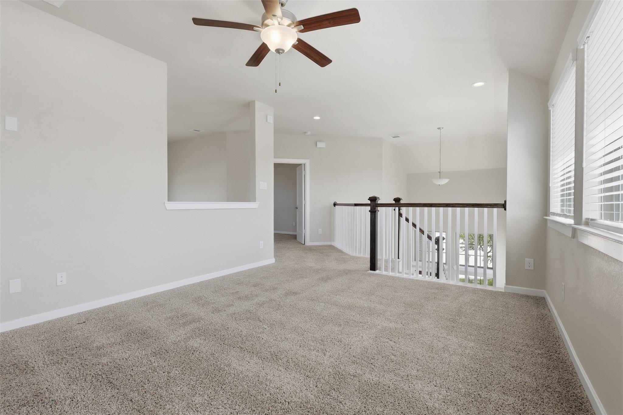 Empty upstairs loft with carpet, ceiling fan, bright windows, and stair railing.