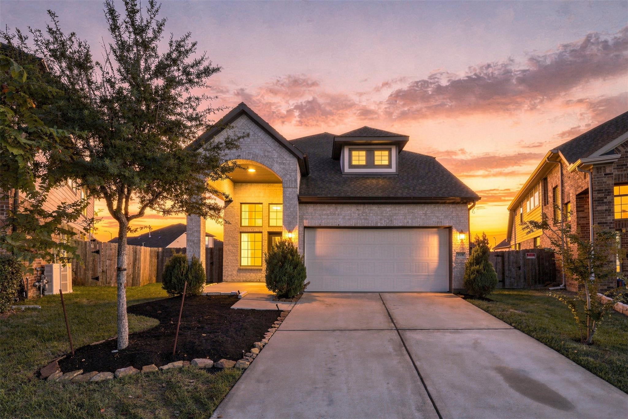 Two-story brick home with lit windows and a wide driveway at sunset.