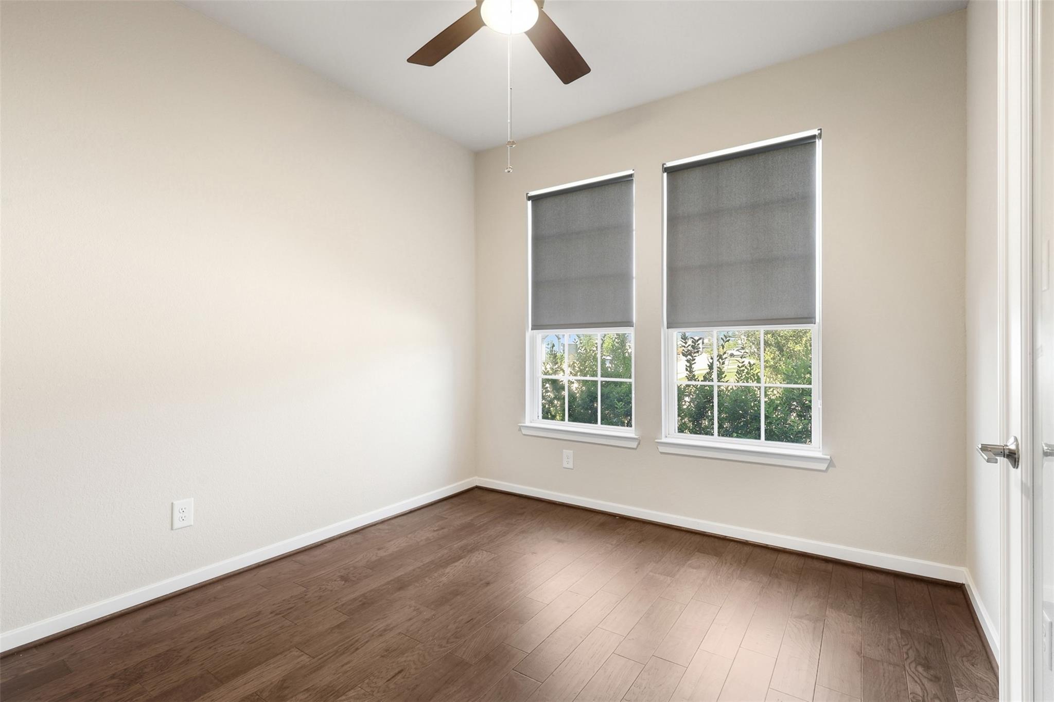 Empty bedroom with dark wood flooring, a ceiling fan, and two shaded windows.