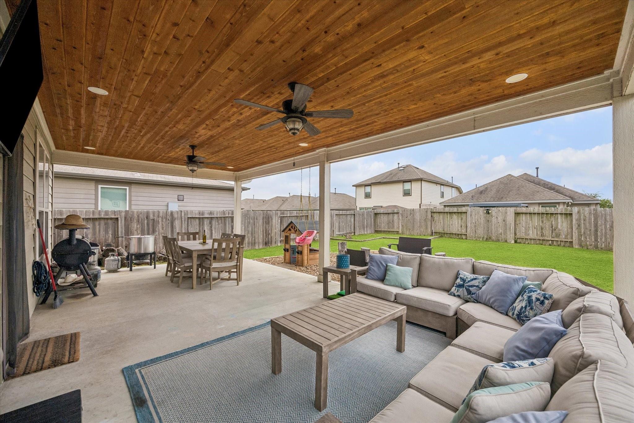 Covered patio with a wood ceiling, outdoor dining set, and sectional seating beside a fenced lawn.
