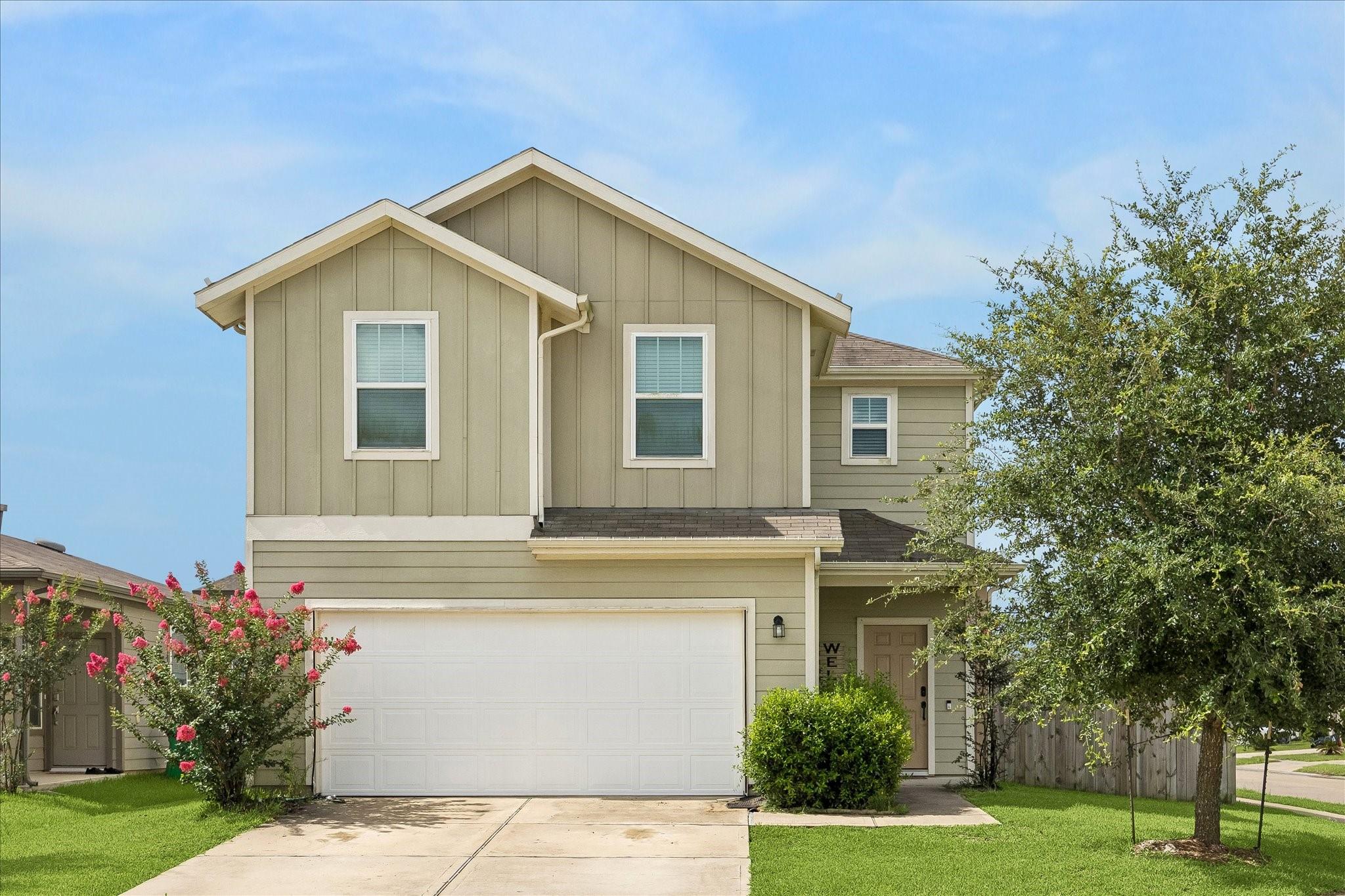 Two-story beige house with a double garage, flowering shrubs, and a large tree in front.