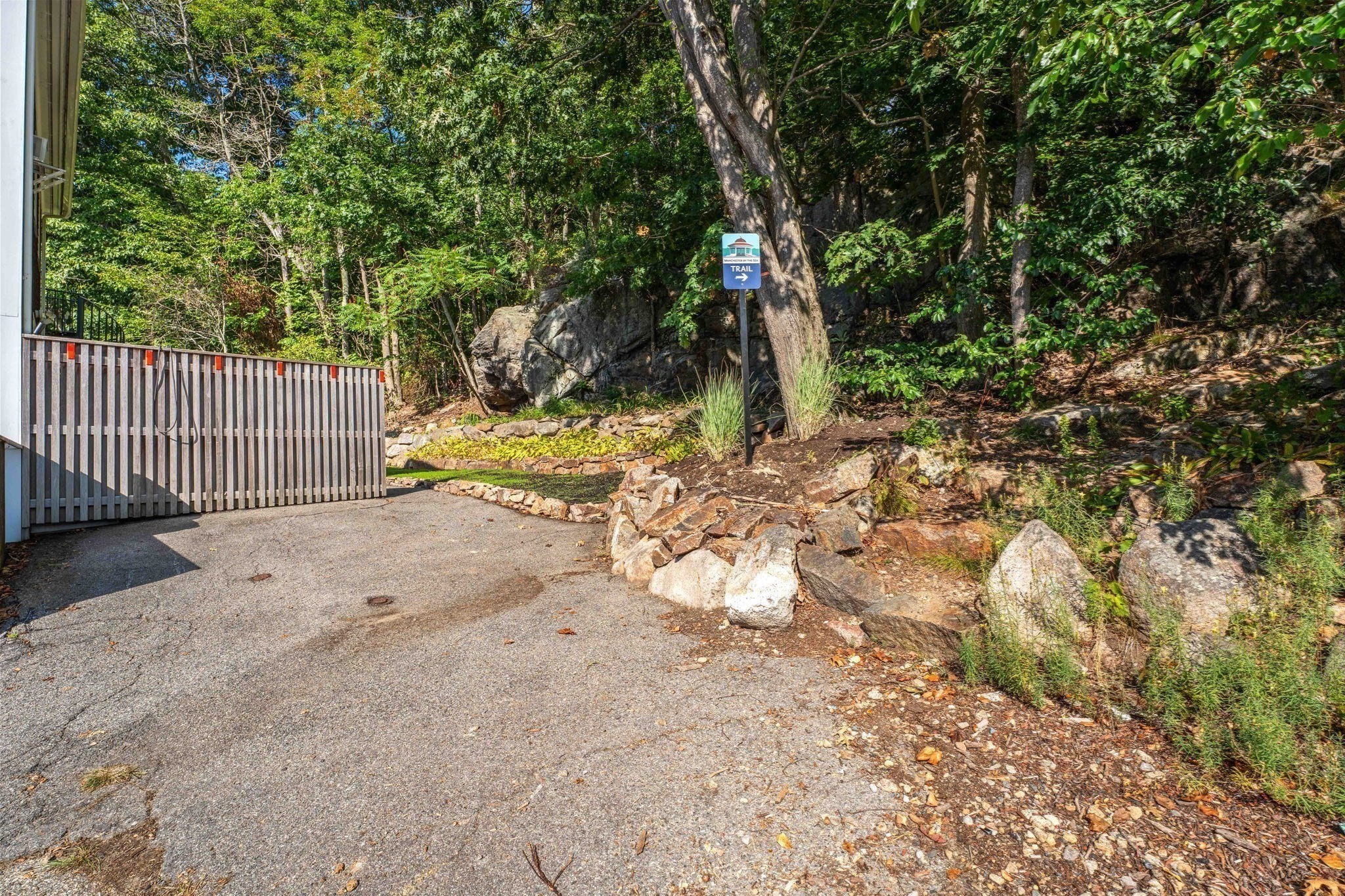 Paved driveway beside wooded landscaping, with stone borders and a trail sign.