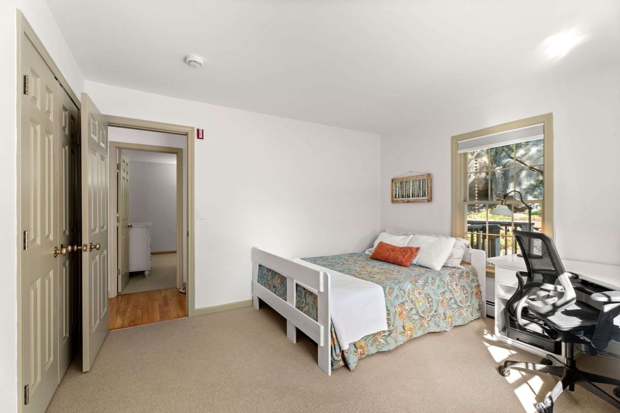 Furnished bedroom with a floral bedspread, desk, and large window overlooking rocks and trees.