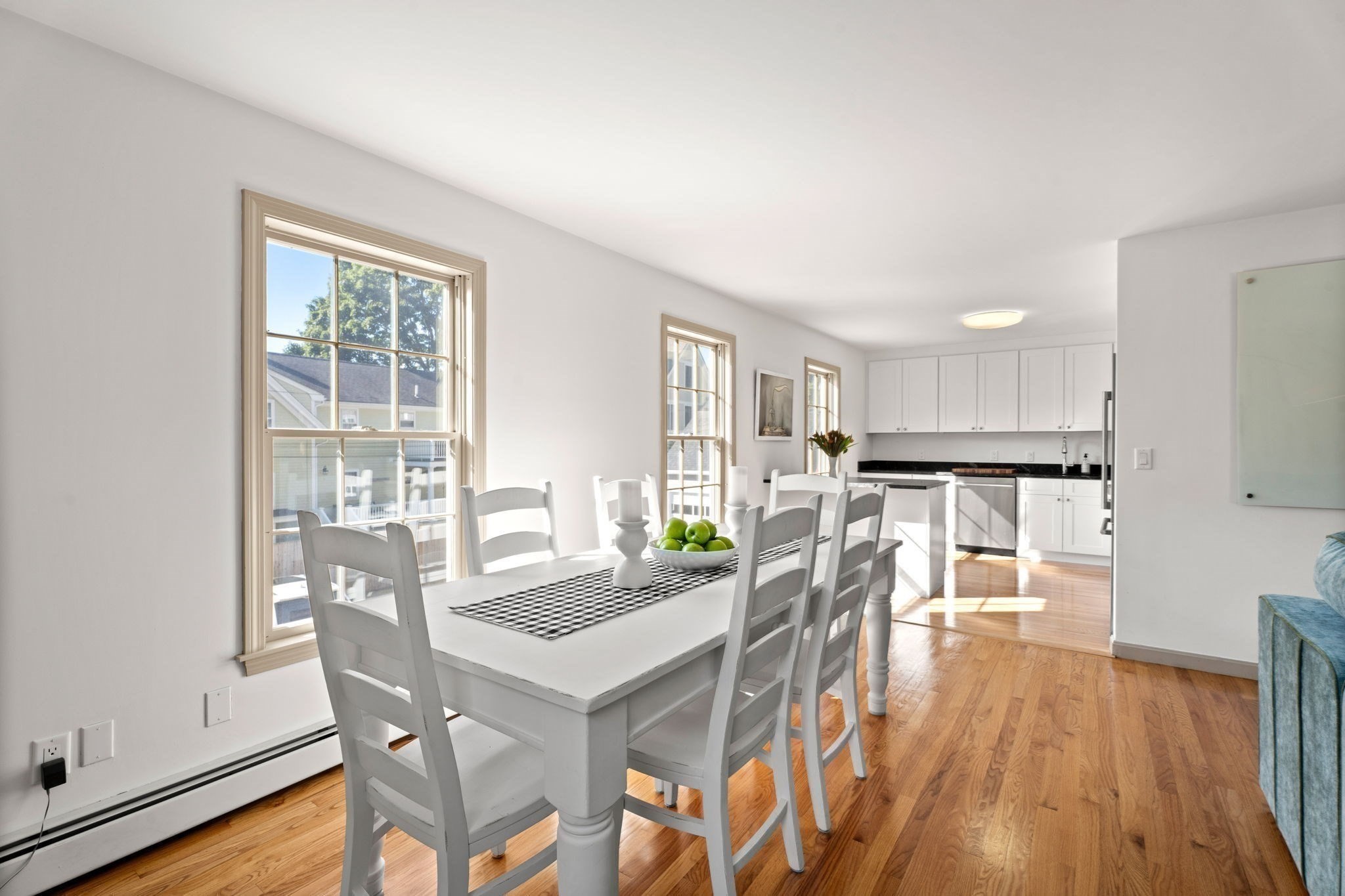 Dining area with a white table beside large windows, opening into a bright kitchen with hardwood floors.