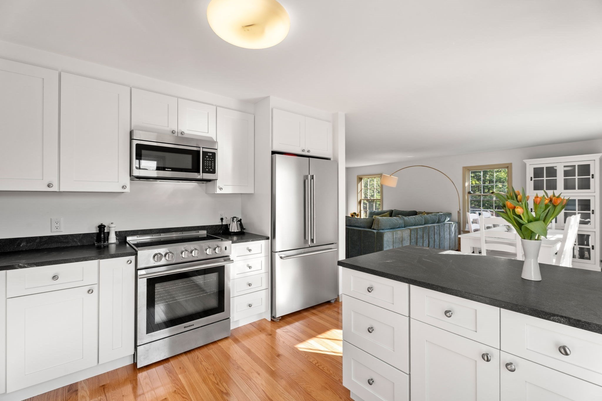 Bright white kitchen with stainless steel appliances and a black island, opening to a living and dining area.
