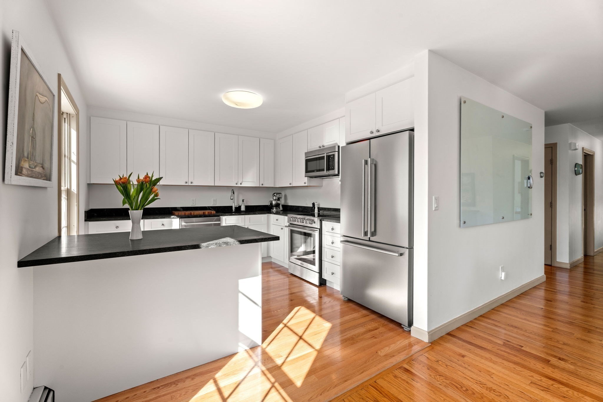 Bright kitchen with white cabinets, black countertops, stainless steel appliances, and sunlight on hardwood floors.
