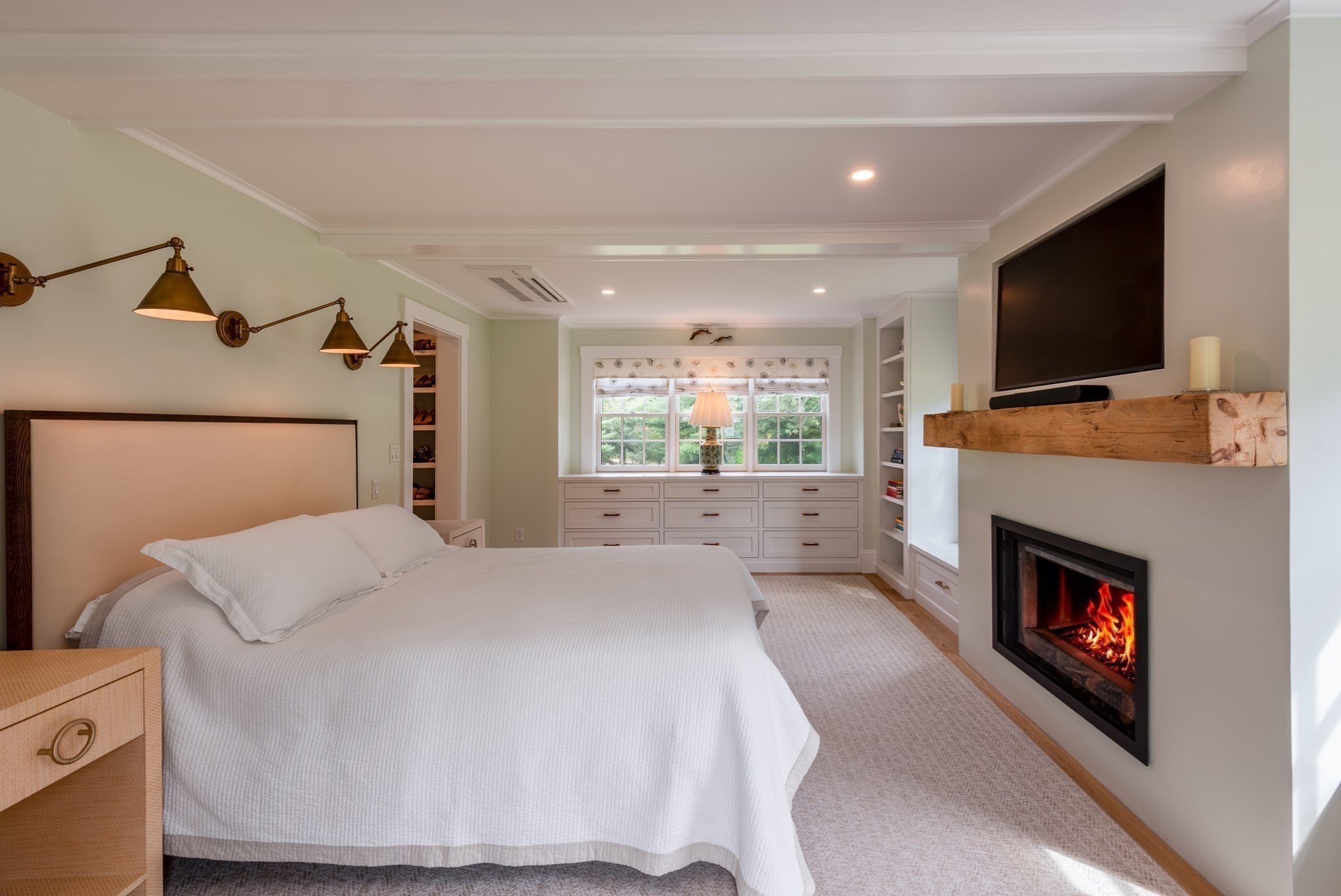 Bedroom with a white bed, built-in window seat drawers, and a lit fireplace beneath a television.
