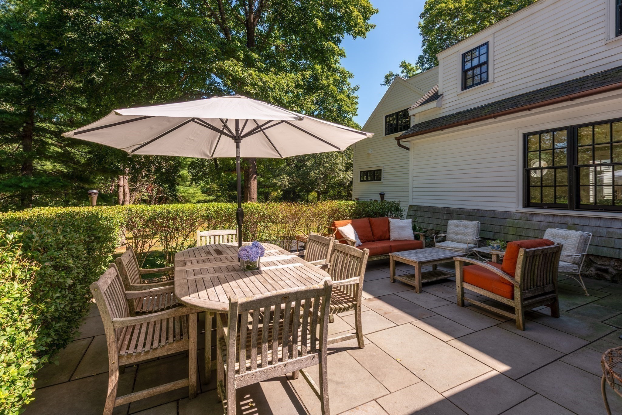 Stone patio with umbrella-covered dining table, lounge seating, and hedges beside the house.