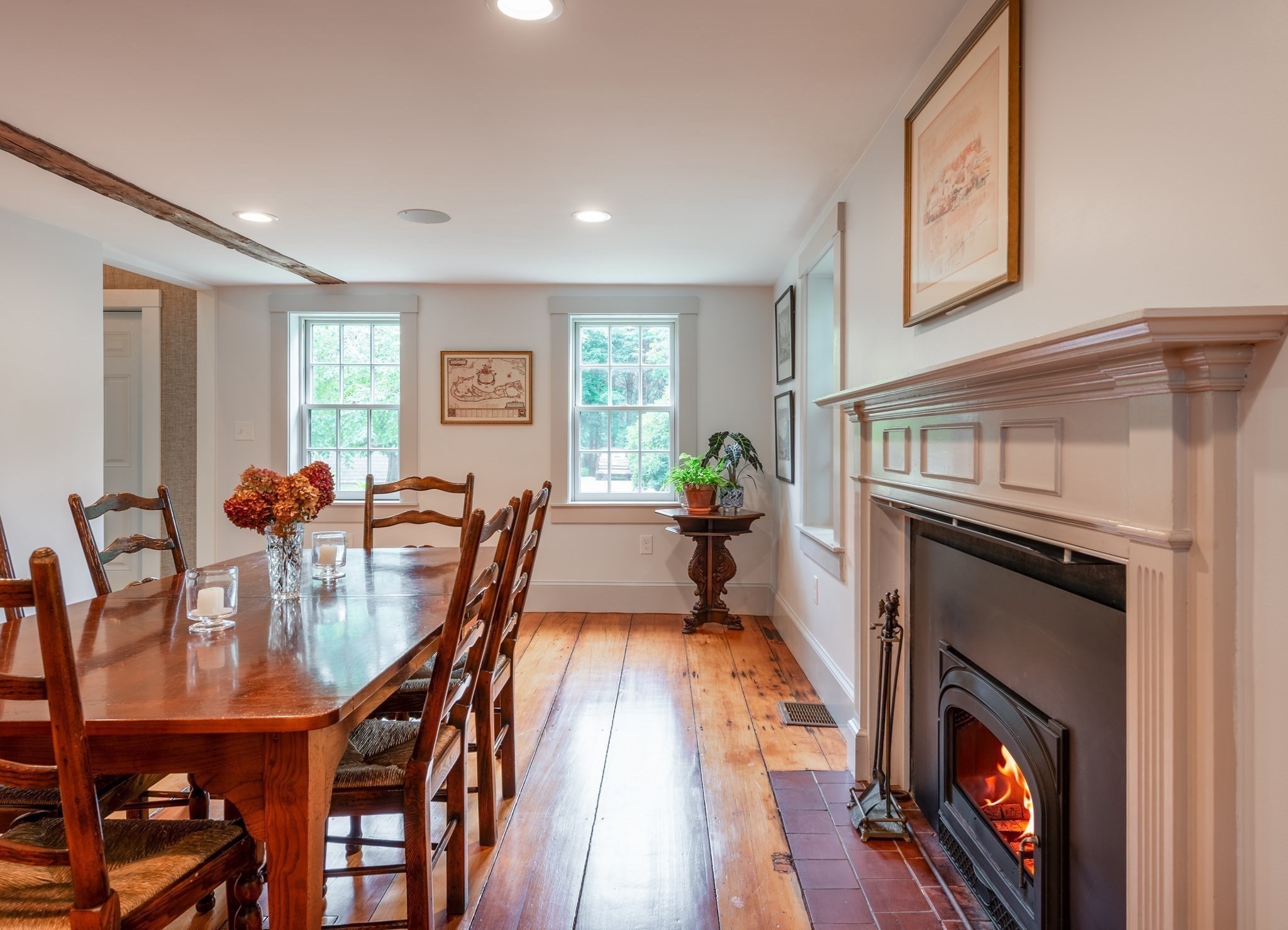 Dining room with a wooden table, wide-plank floors, and a lit fireplace beneath a paneled mantel.