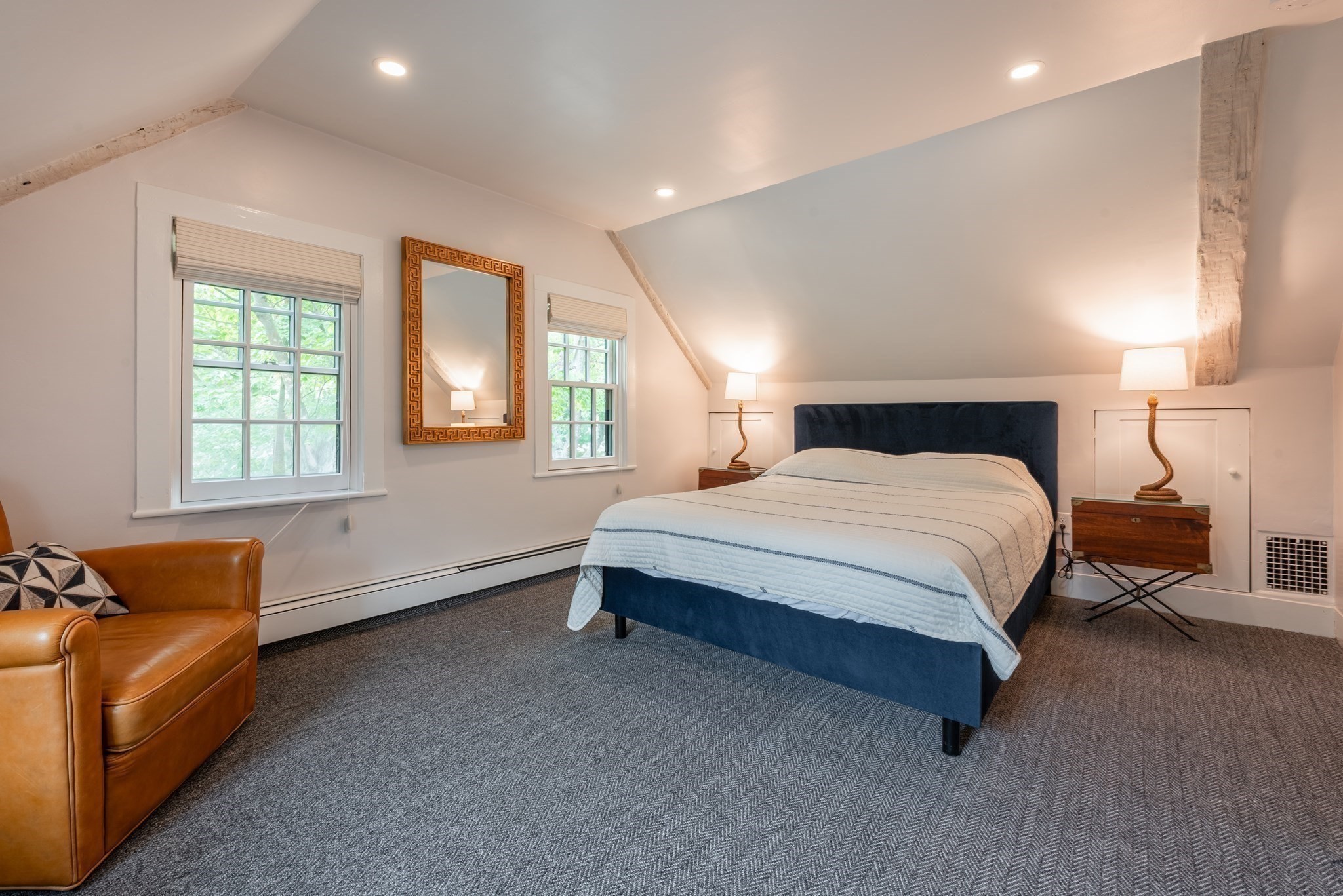 Attic bedroom with a blue bed, exposed beams, two windows, and a tan leather chair.