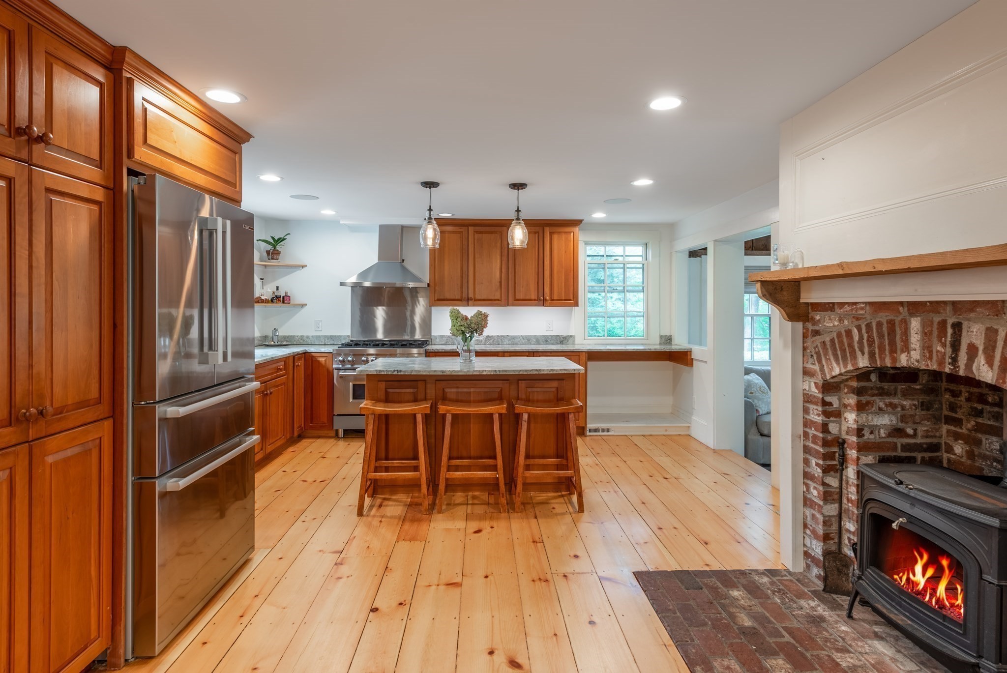 Kitchen with warm wood cabinets, a central island, wide-plank floors, and a brick fireplace with a lit stove.