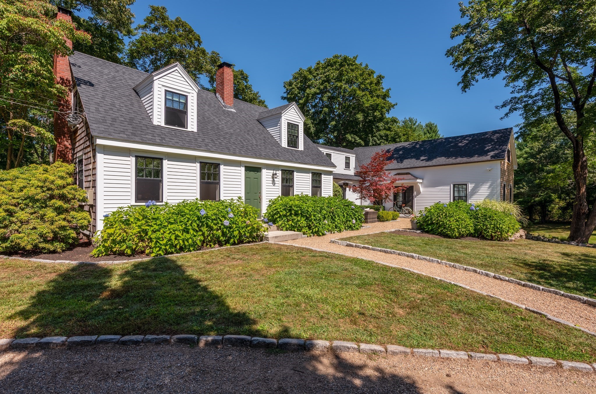 White clapboard house with dormers, brick chimneys, a green door, and hydrangeas along the front.