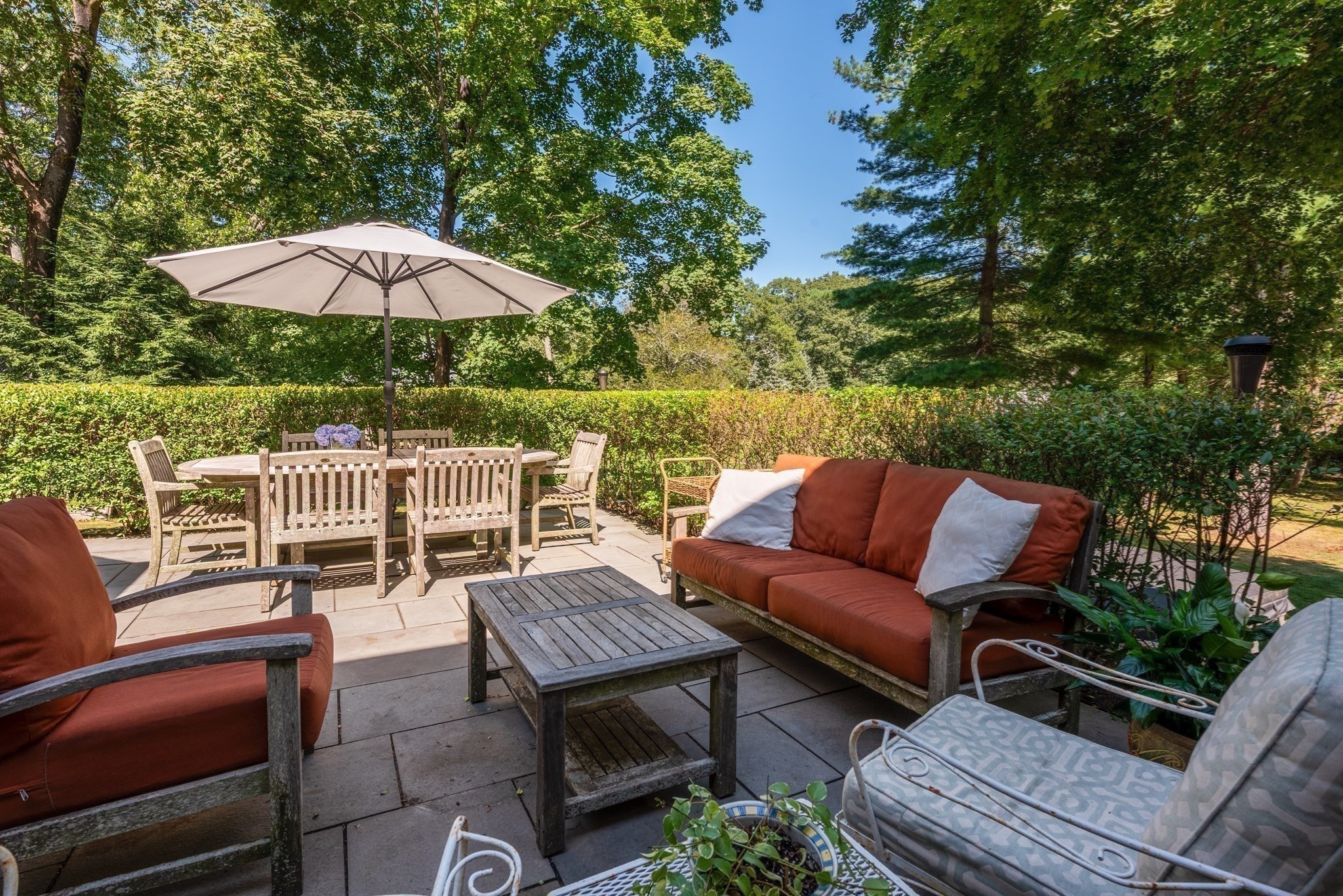 Stone patio with dining set and umbrella, lounge seating, and leafy hedges.
