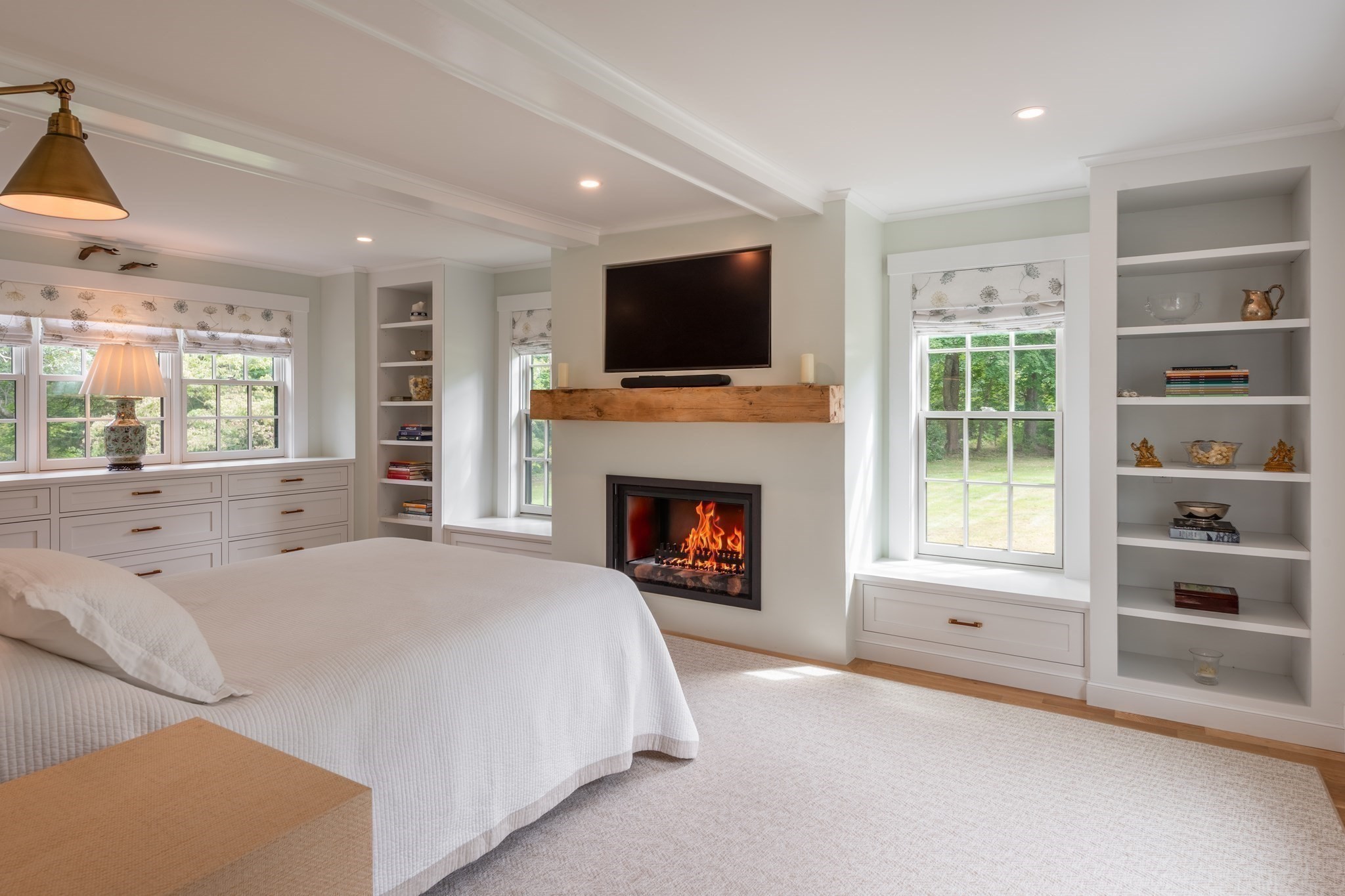 Bedroom with a lit fireplace, built-in shelving, and large windows overlooking greenery.