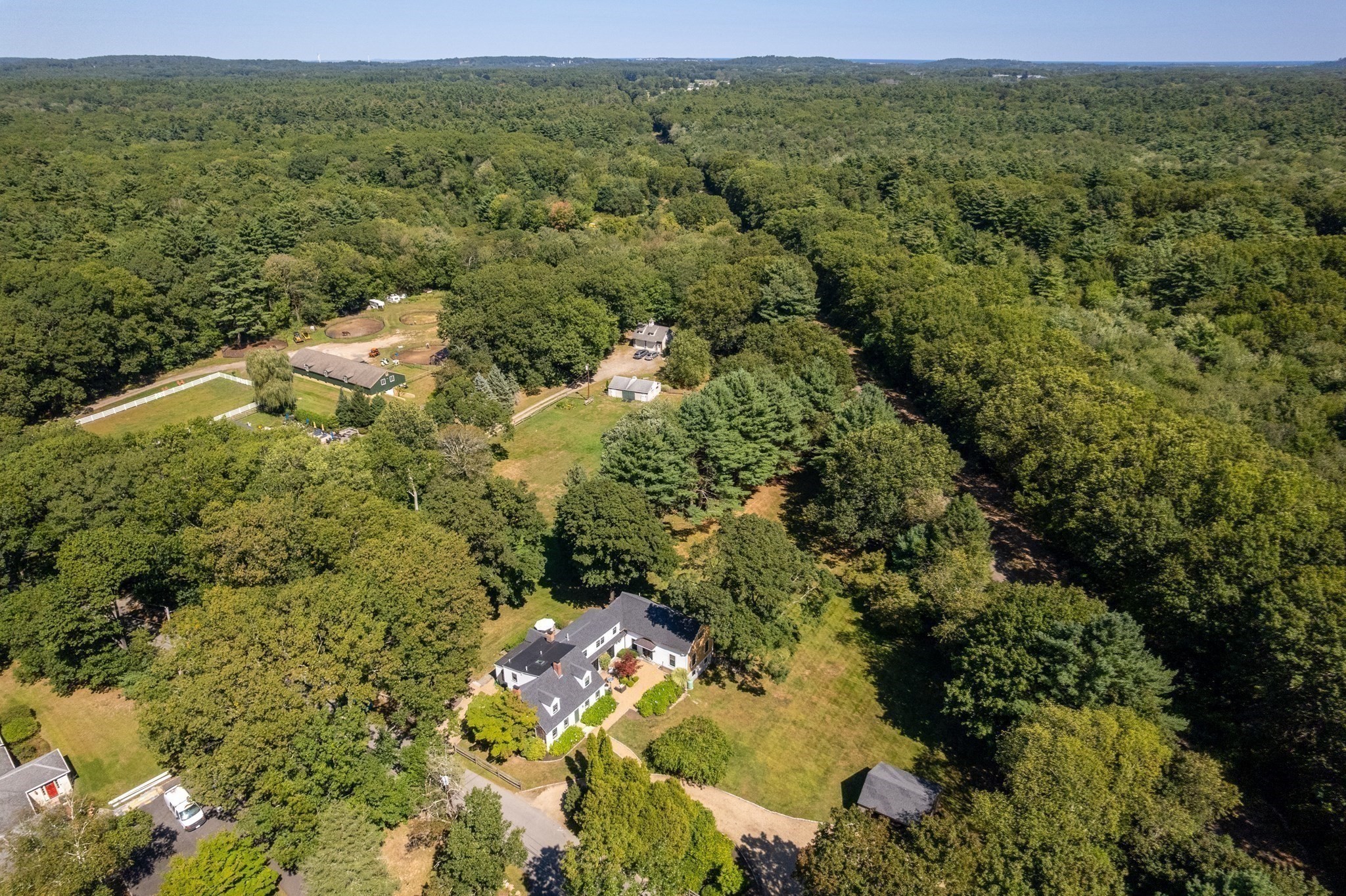 Aerial view of a home surrounded by wooded grounds, lawns, outbuildings, and fenced areas.