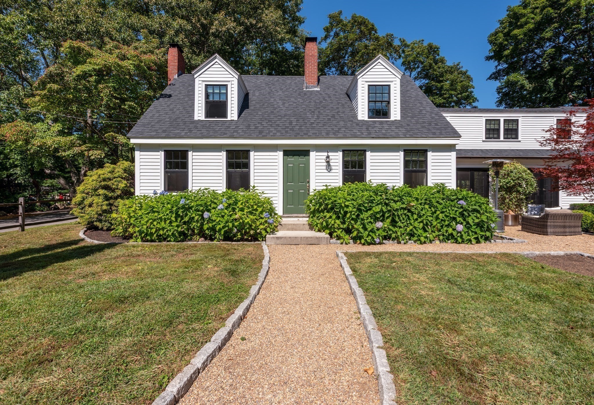 White clapboard house with two dormers and green door, approached by a gravel path through hydrangeas.