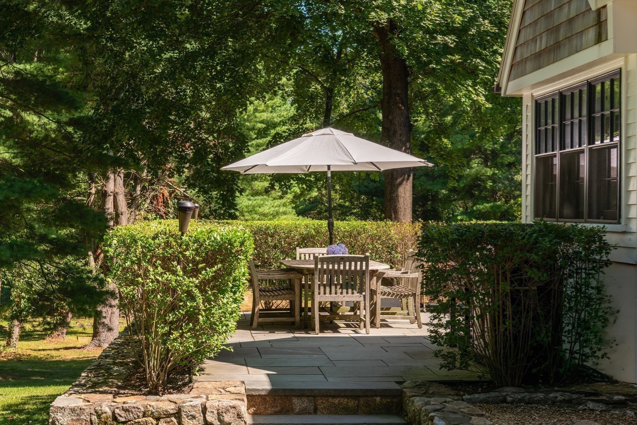 Stone patio with wooden dining set and umbrella, surrounded by hedges and mature trees.