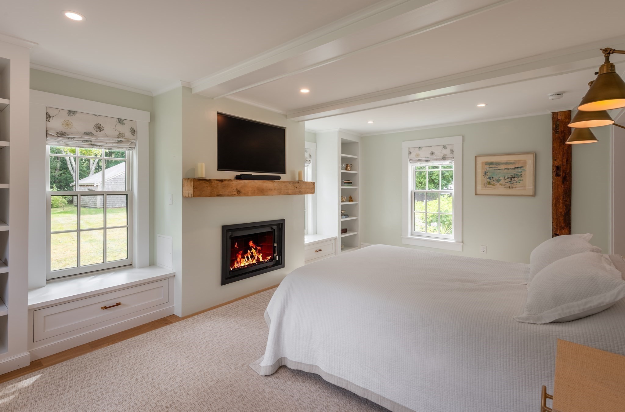 Bedroom with a white bed, built-in window seats, and a lit fireplace with a wood mantel.
