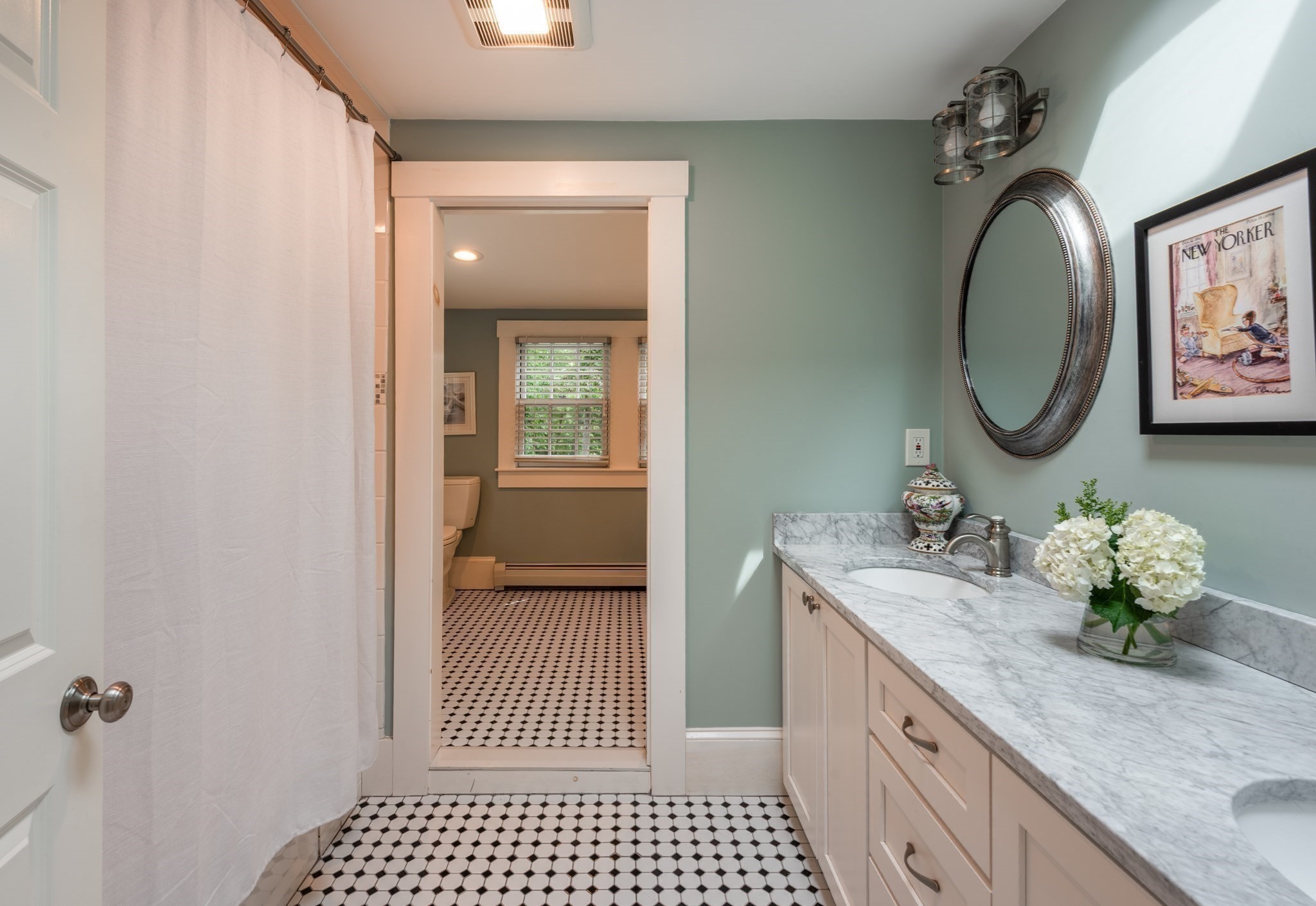 Bathroom with marble double vanity and geometric tile floor, looking through to a toilet area with a window.