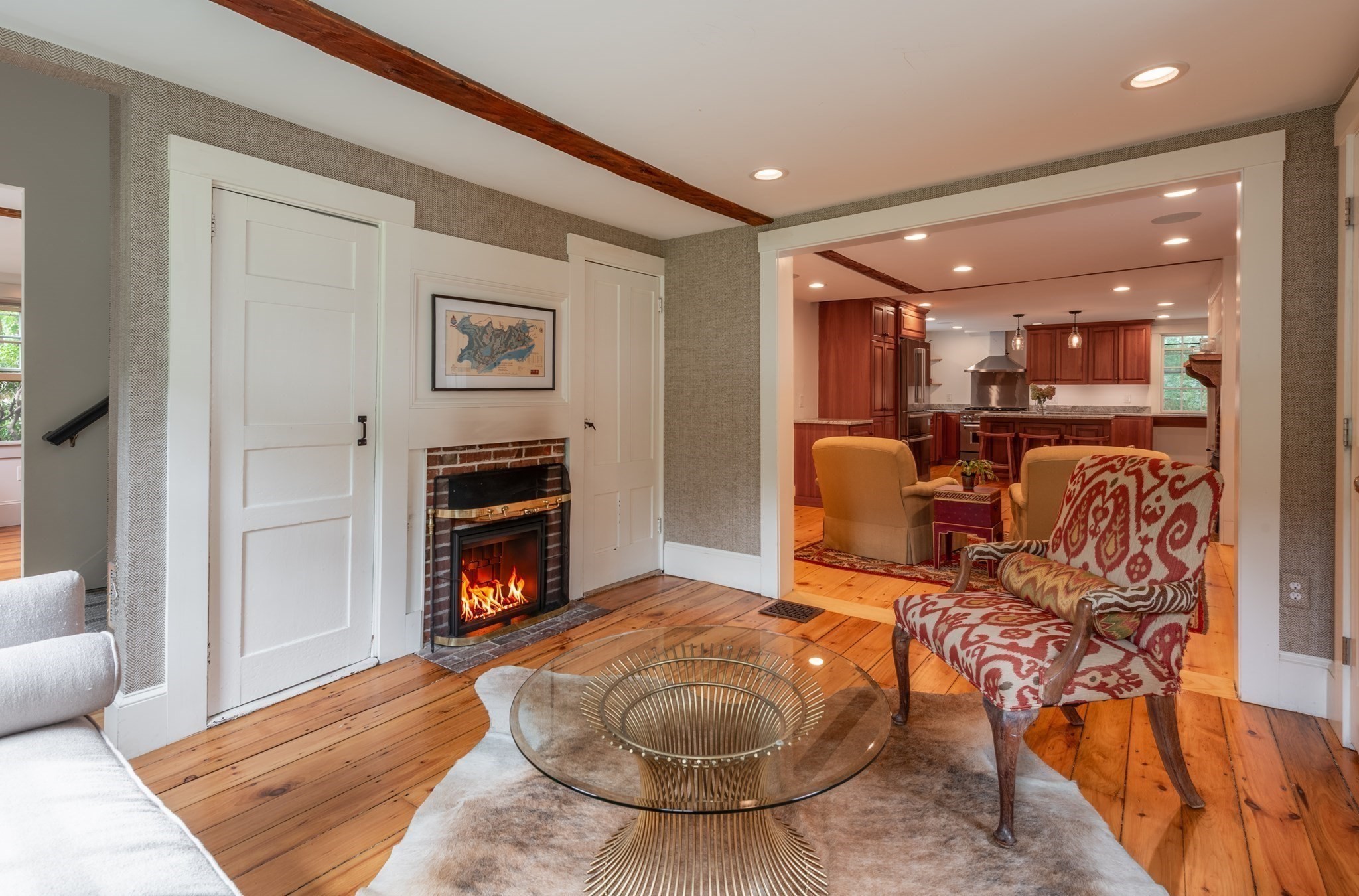 Living room with a brick fireplace, patterned armchair, and wood floors opening to a kitchen.